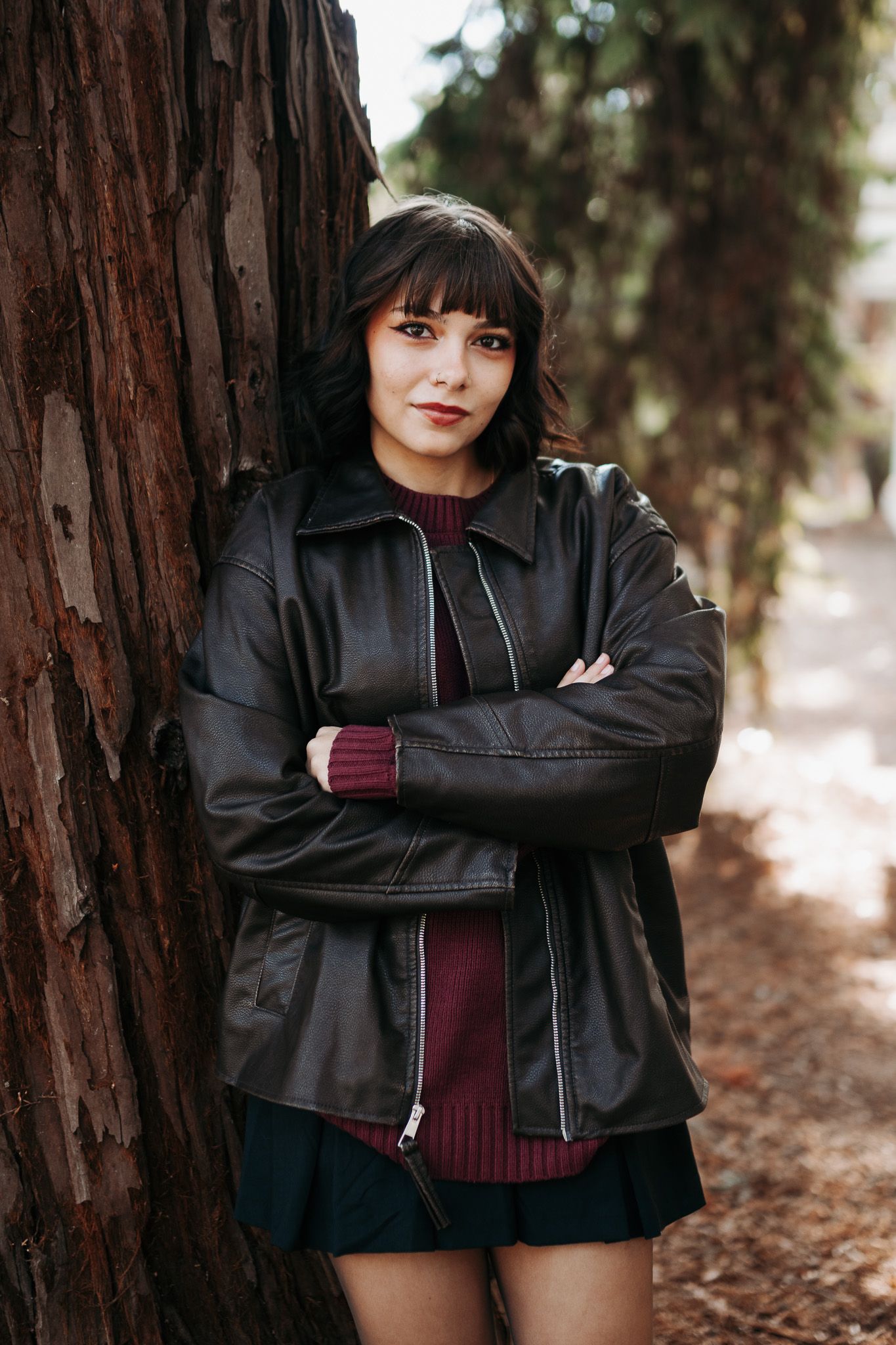 Woman in black leather jacket and burgundy sweater leans against large tree trunk on woodland path.