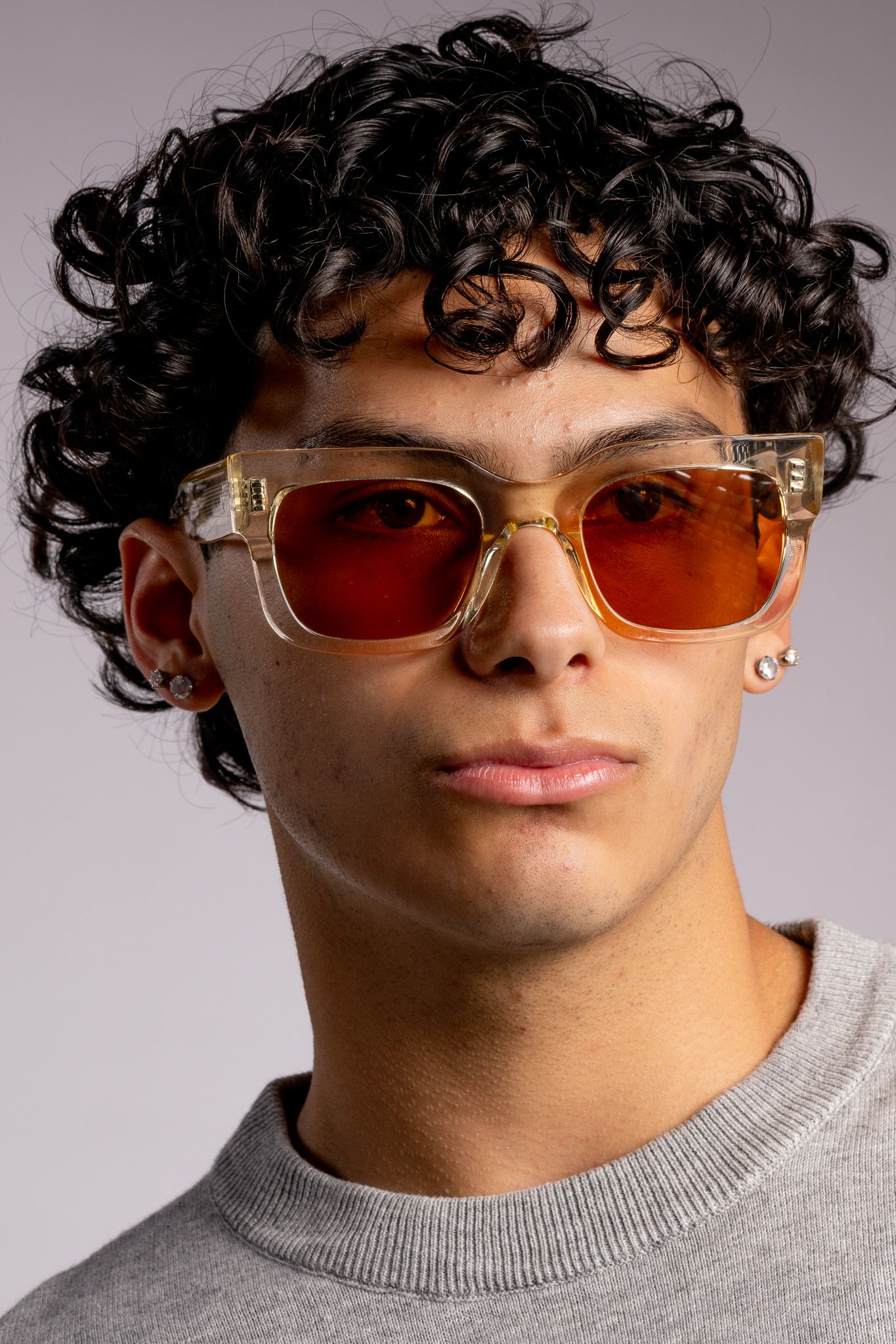 A fashion model wearing orange-tinted clear frame sunglasses and a grey sweater poses against a grey backdrop.