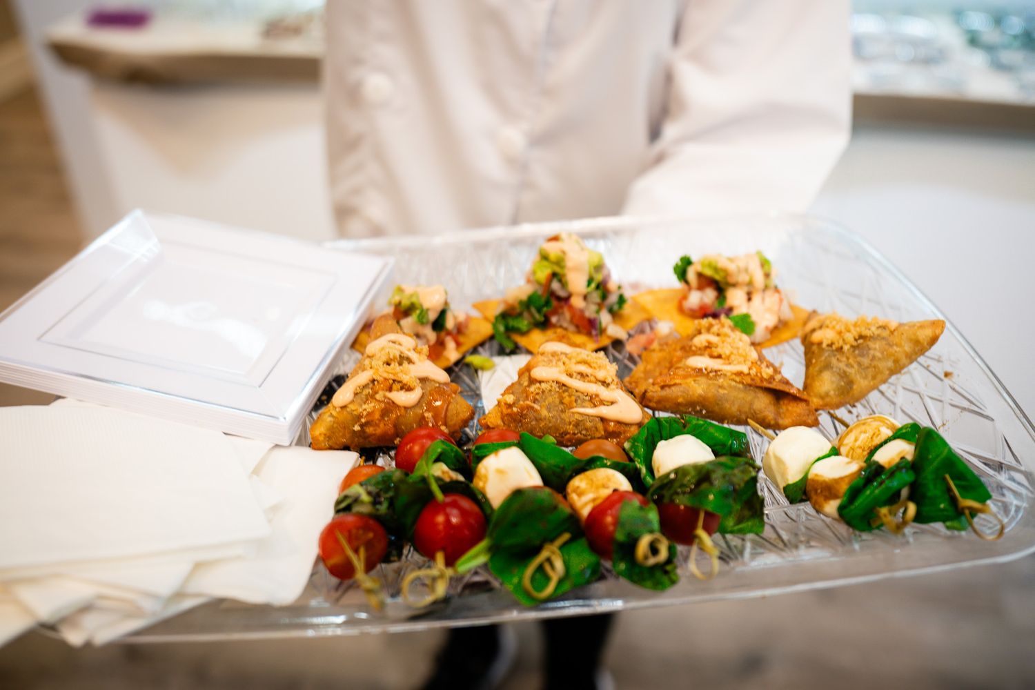 Catering tray with sandwiches, tortilla chips, and fresh vegetables arranged on white paper with garnish.