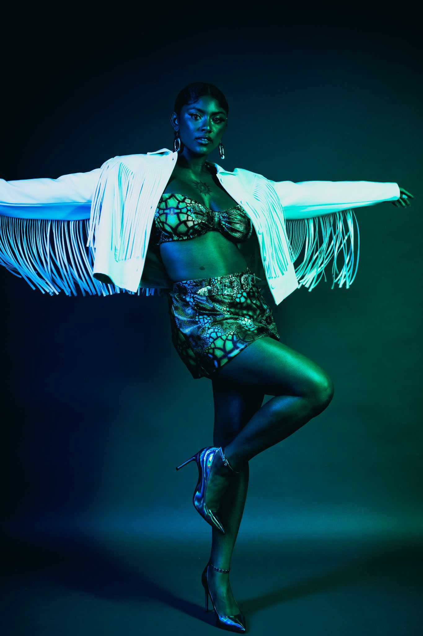 Dancer in white fringed jacket and patterned swimwear strikes dramatic pose against dark background with blue-green lighting.