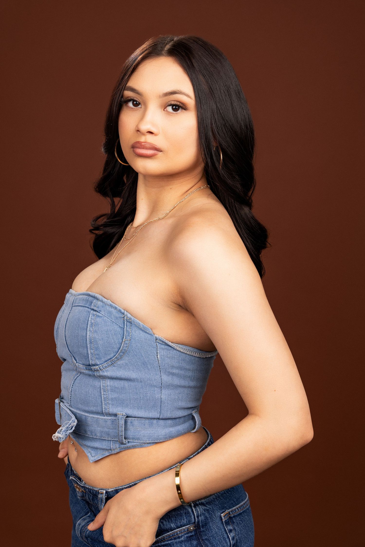 Studio portrait of someone wearing a strapless denim crop top and jeans against a brown background.