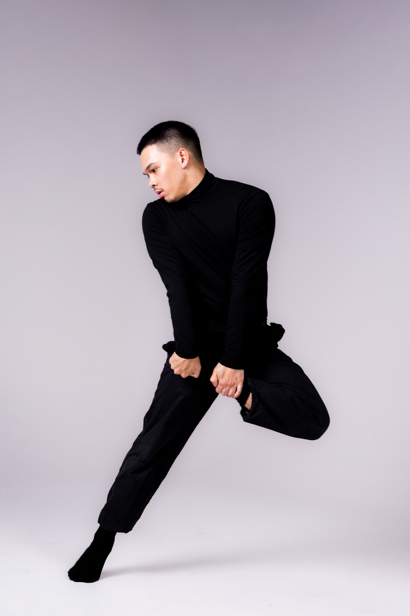 Performer in all black attire strikes a dynamic dance pose against a light gray studio backdrop.