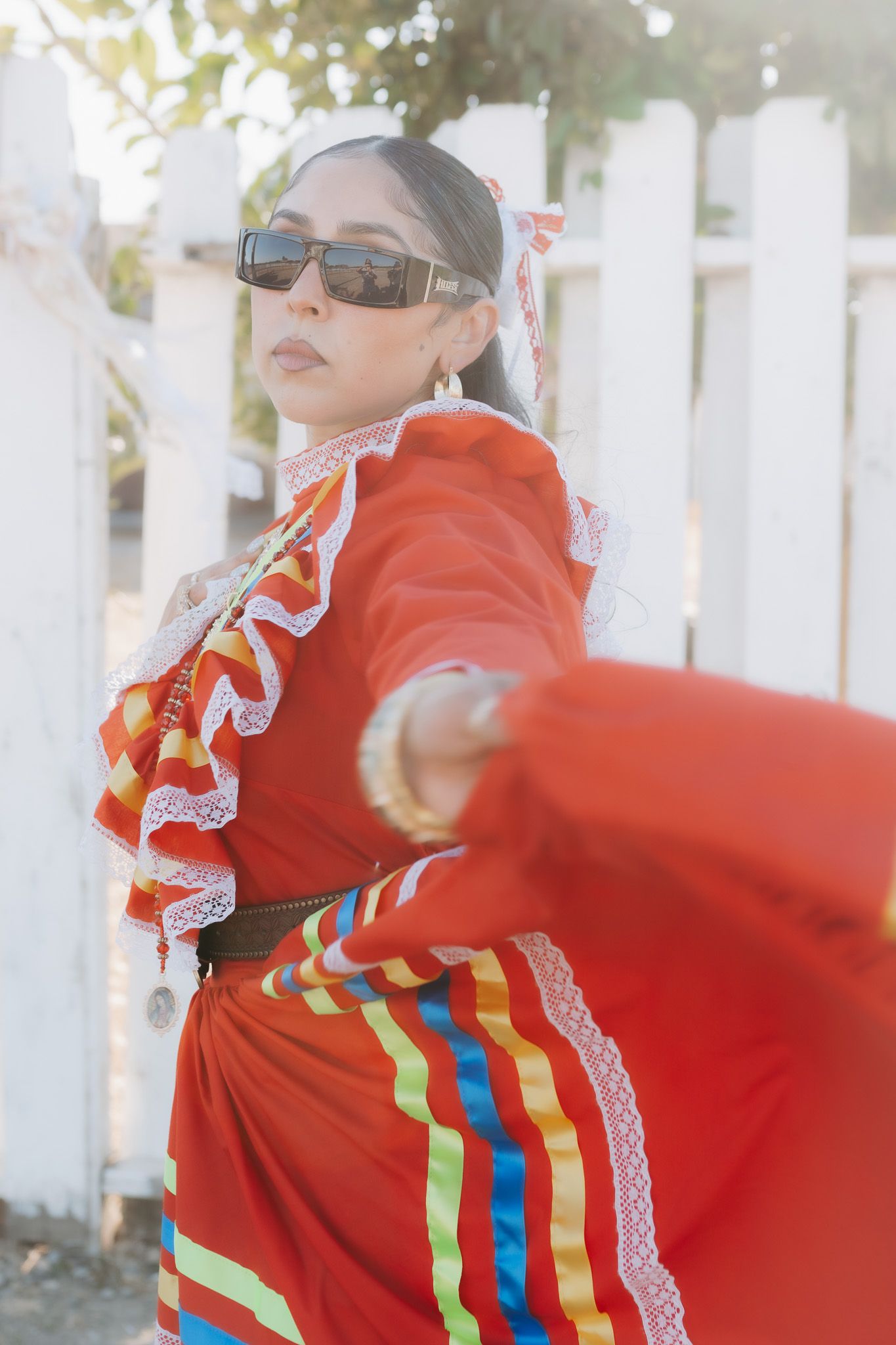 A vibrant red traditional Mexican dress with colorful ribbons and lace trim is worn against a white picket fence background in sunlight.