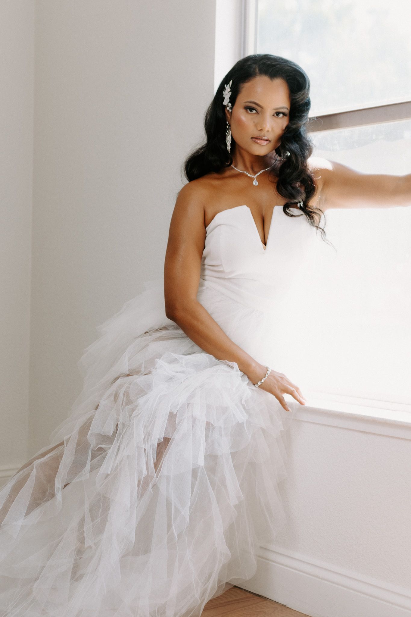 Beautiful bride in a white strapless wedding gown with ruffled tulle skirt poses elegantly by window in natural light.