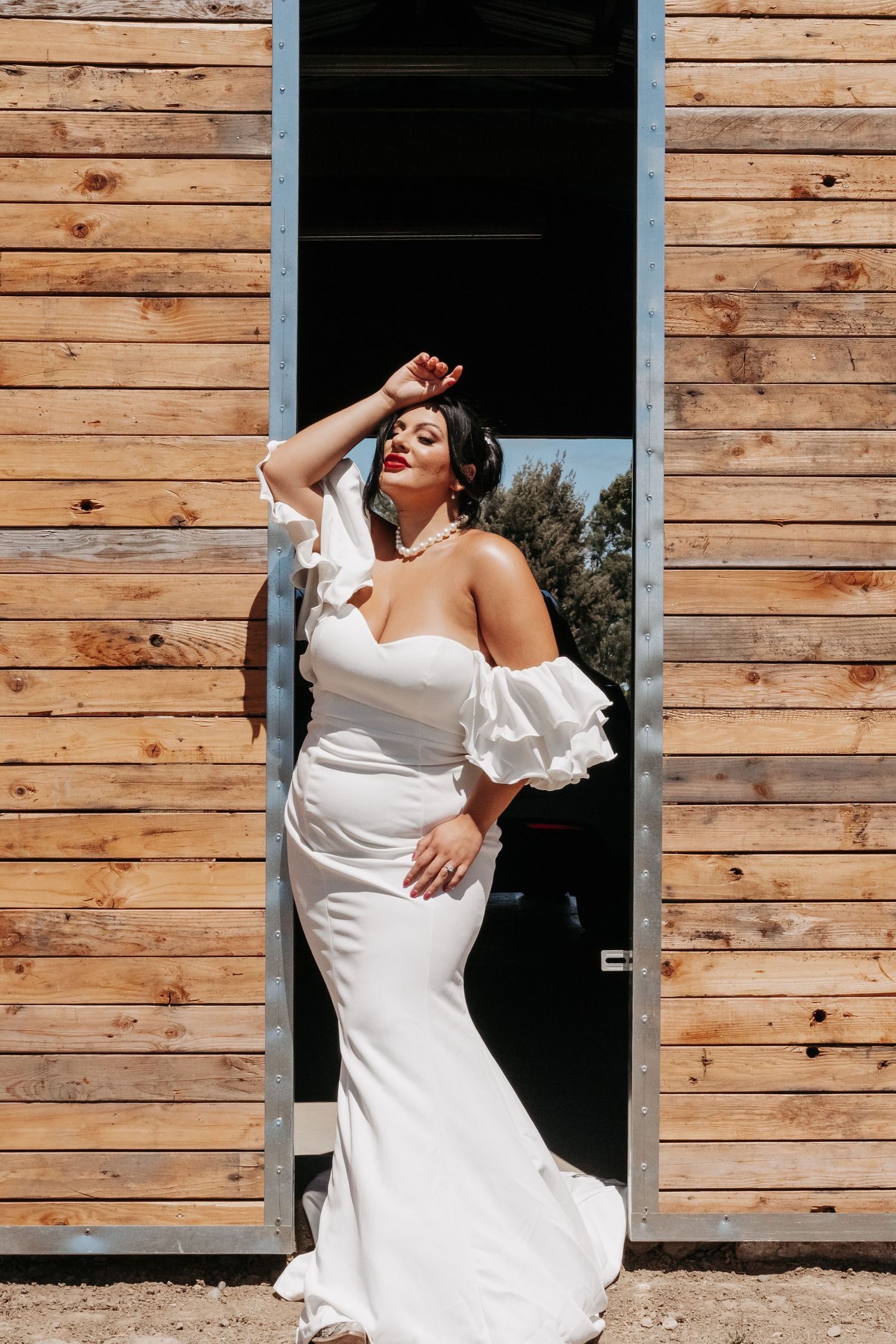 A bride in an elegant off-shoulder white gown poses dramatically against a rustic wooden wall with a blue doorway.