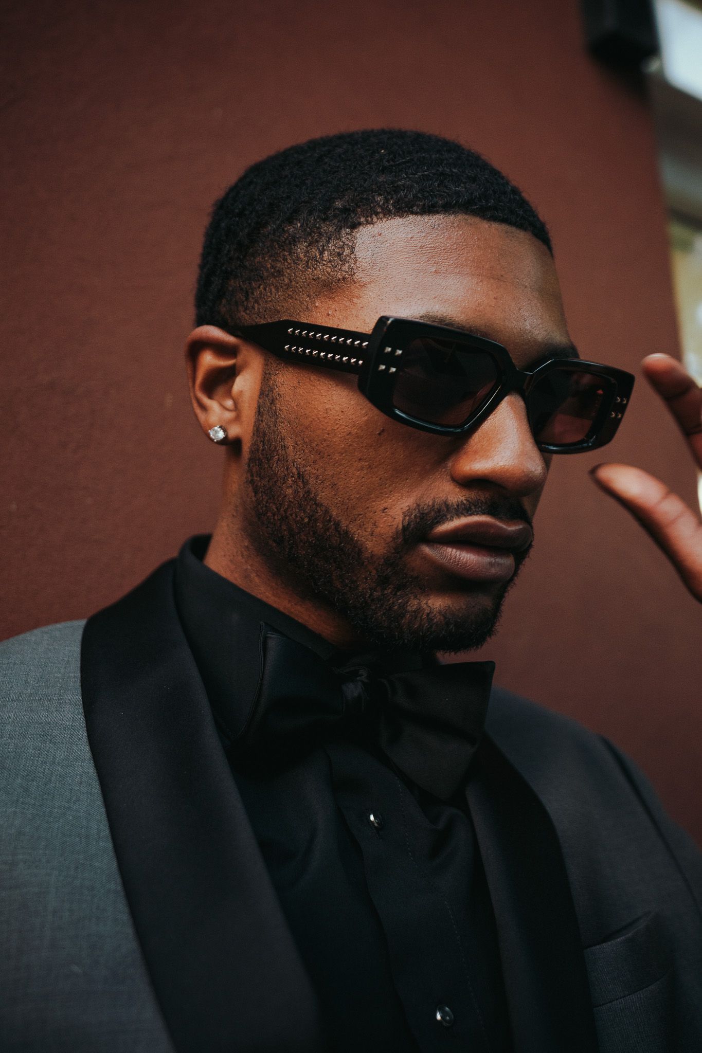 Stylish portrait of person wearing black suit, bow tie and designer sunglasses against red wall.