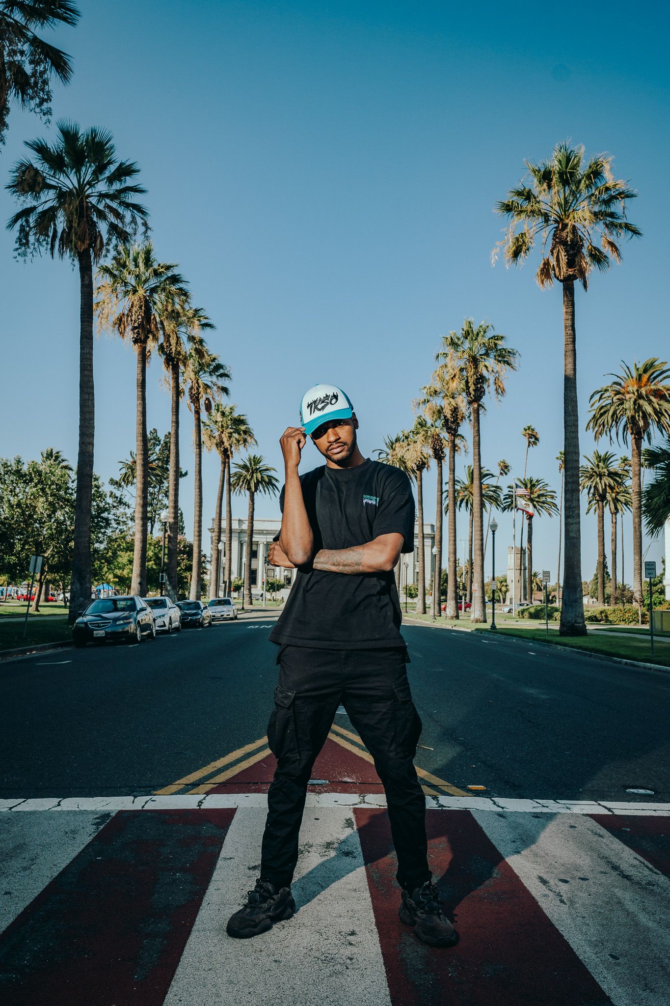 Urban street photography with palm trees lining a road while someone poses on a crosswalk wearing black and a blue cap.