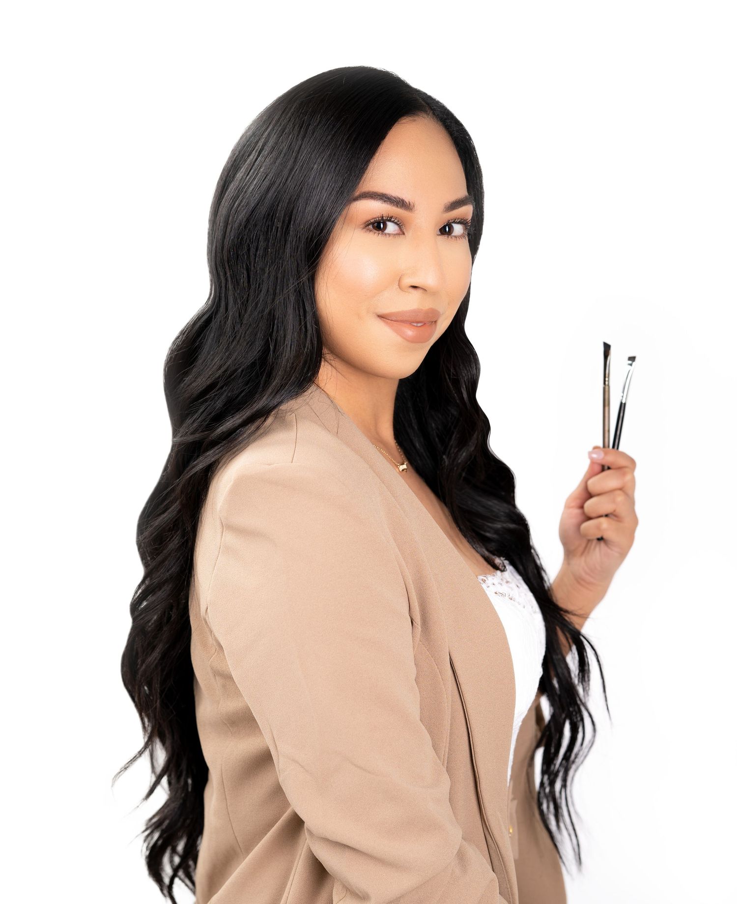 Professional beauty services provider in beige blazer holding styling tools stands against white background with long wavy dark hair.