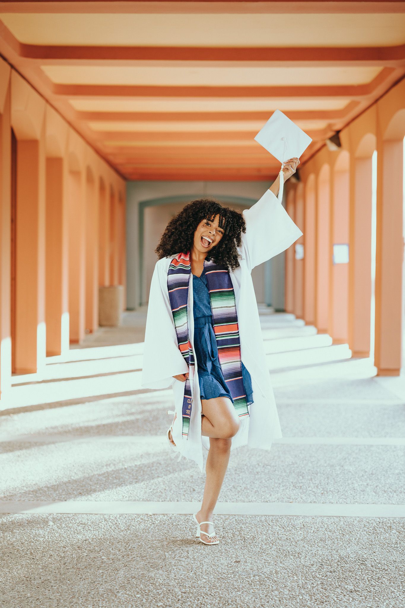 Graduate in white gown and colorful dress celebrates with papers flying in peach-colored colonnade hallway.