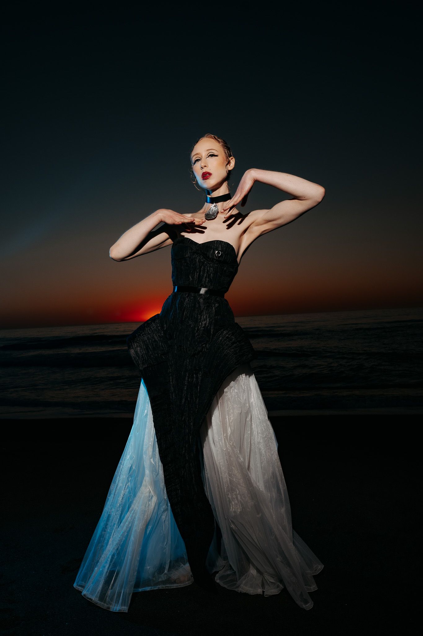 A dramatic fashion photo of a model in a black and white gown posing expressively against a sunset sky.