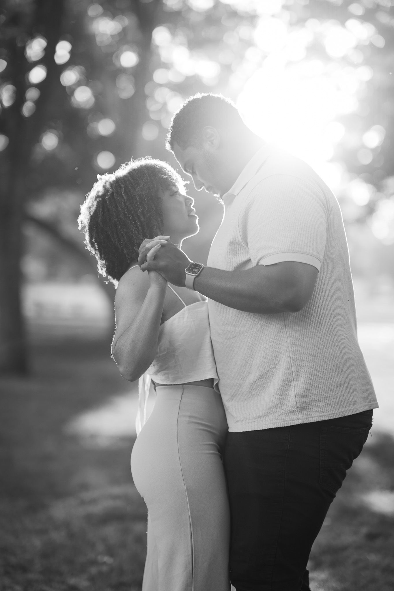 A romantic black and white outdoor photo of a couple sharing a tender moment in backlit sunlight with bokeh effects.
