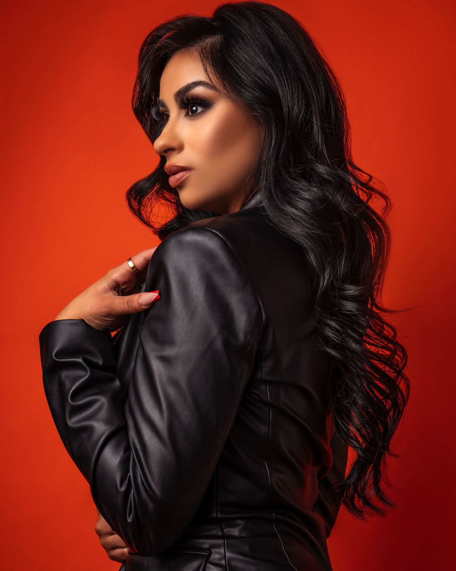 A model wearing a black leather jacket poses against a vibrant red background, with long dark wavy hair cascading down.