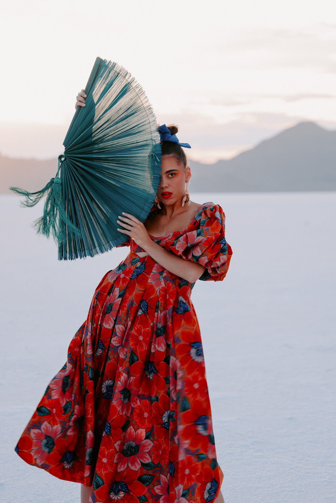 Figure in a vibrant red floral dress poses with a teal fan against a white salt flat landscape at sunset.