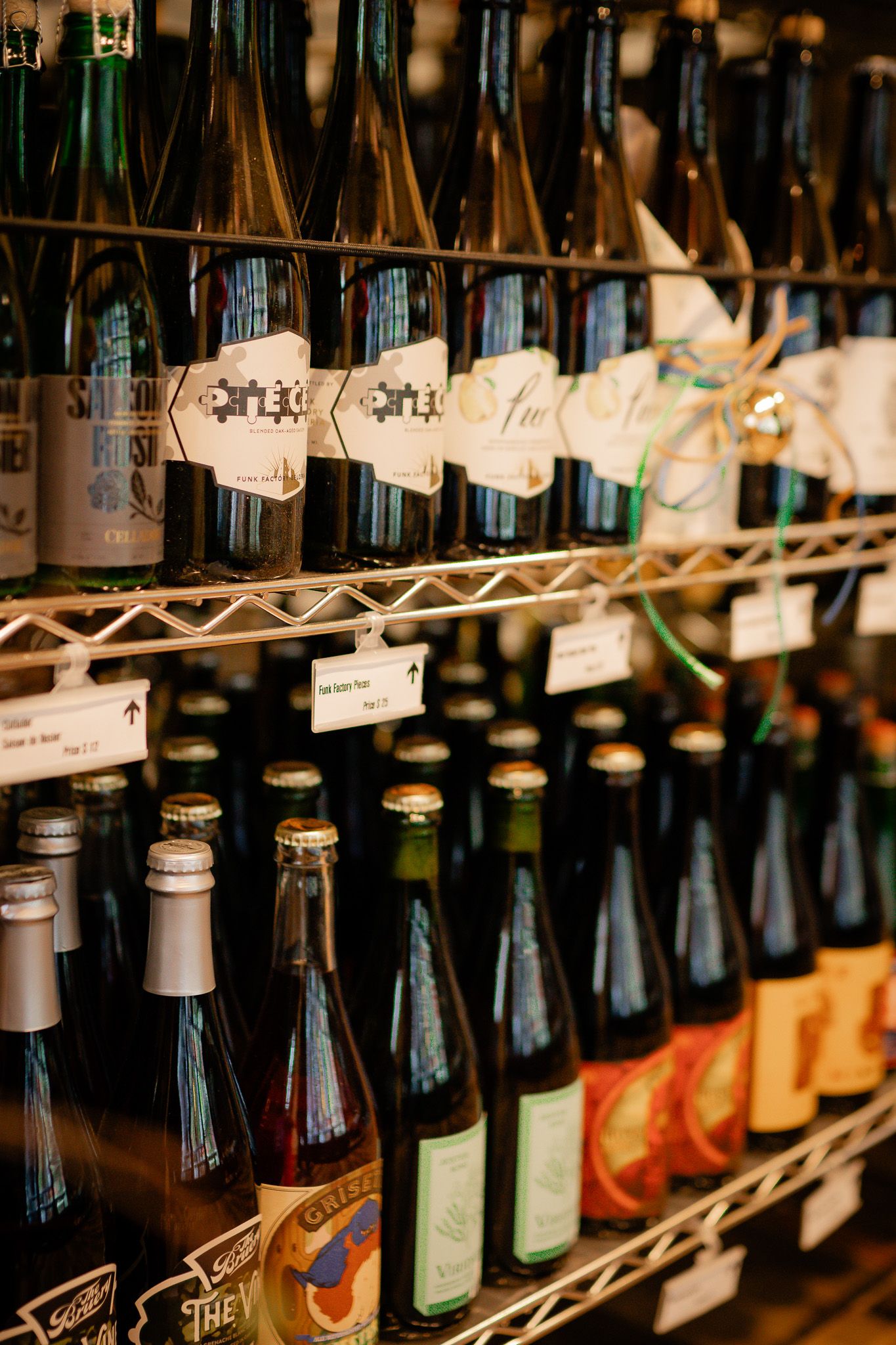Rows of artisanal beer bottles with custom labels displayed on metal wire shelves in a specialty craft beer store.