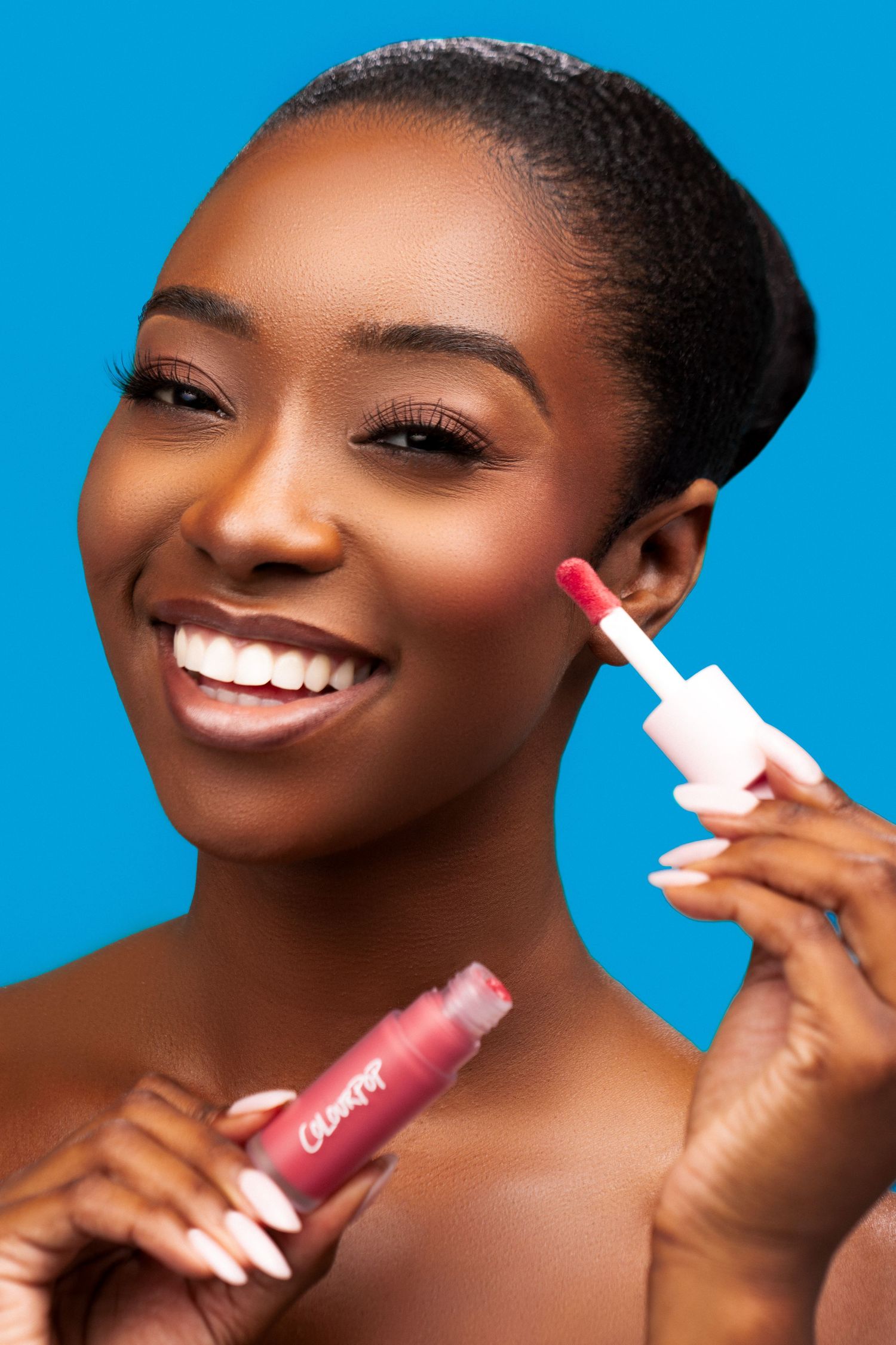 Beauty model smiling while applying pink lip gloss against bright blue background.