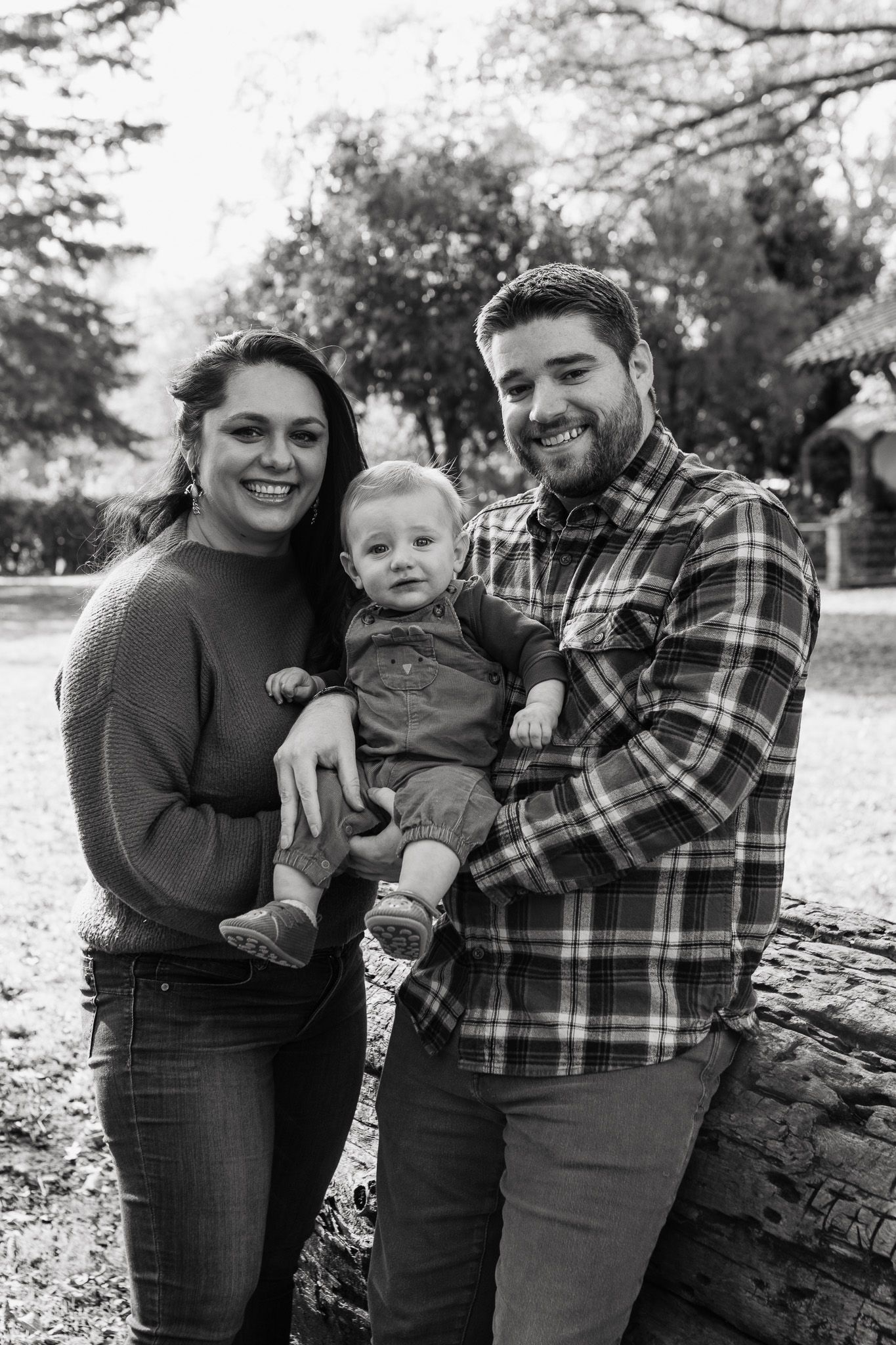 A black and white portrait of a family posing together outdoors in autumn, standing next to a fallen log.
