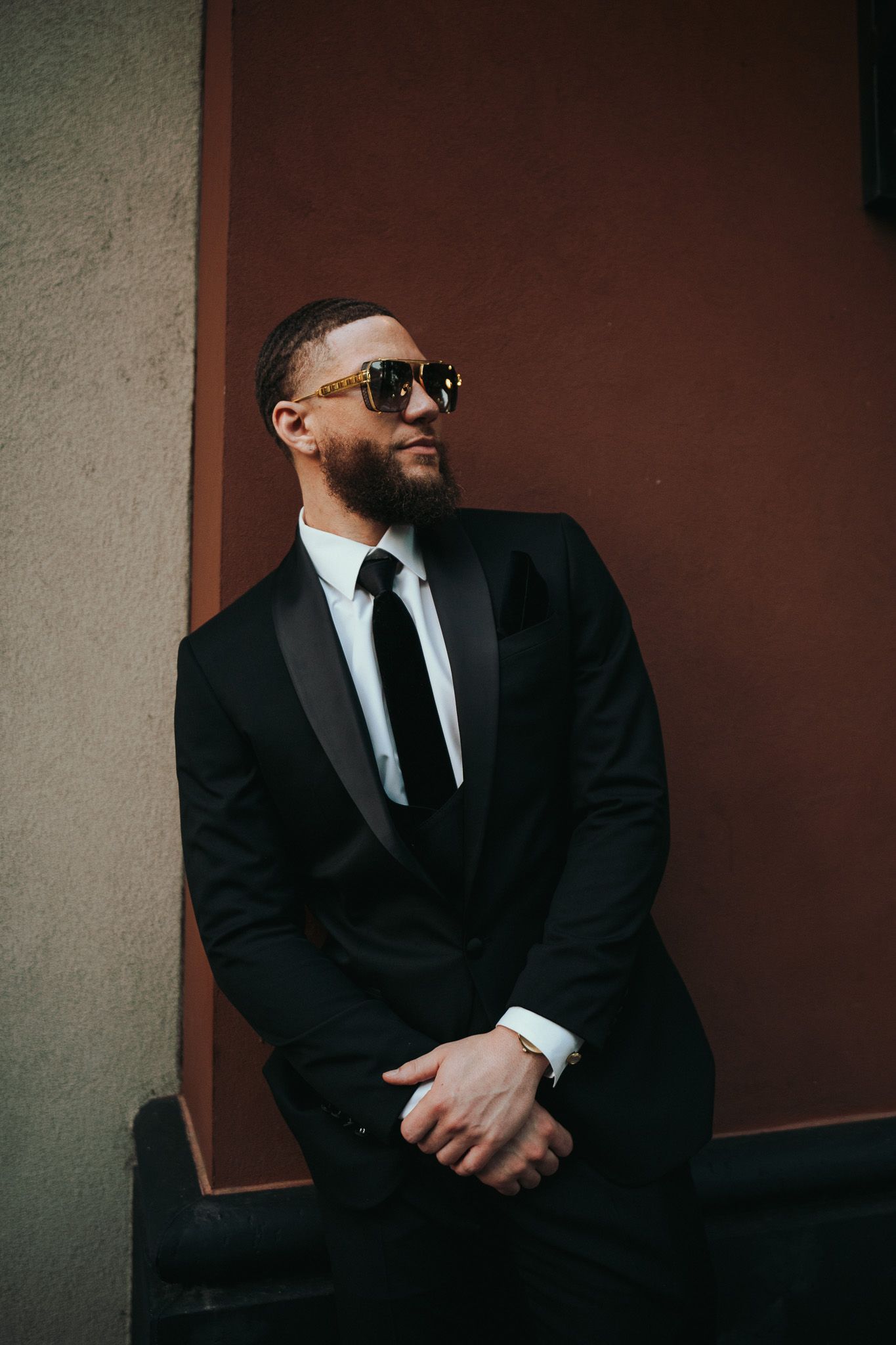 A stylishly dressed man in a black suit and sunglasses poses against a wall in a moody portrait shot.