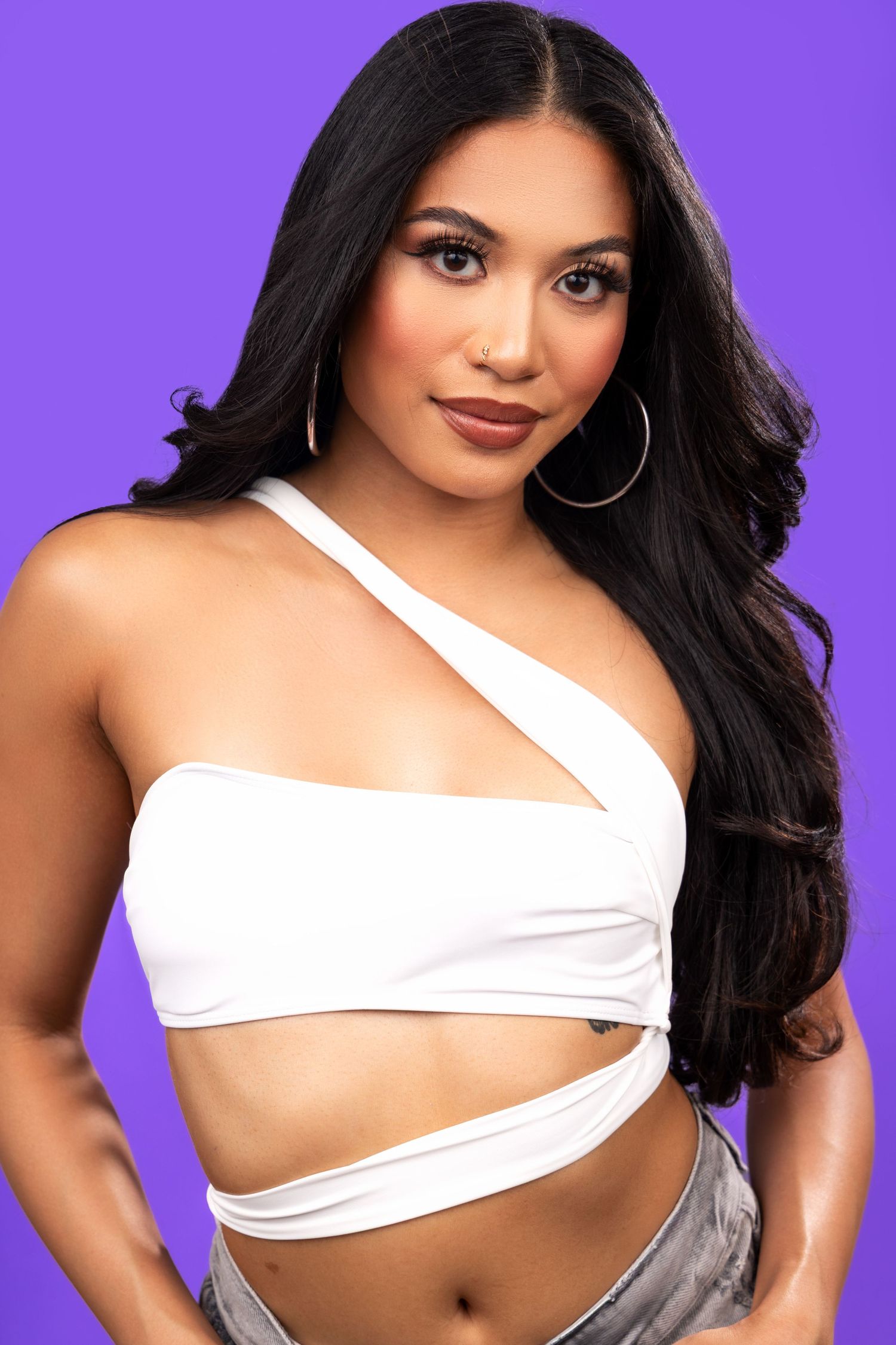 Portrait against purple backdrop shows a glamorous look with long dark hair, white asymmetrical crop top and silver hoop earrings.