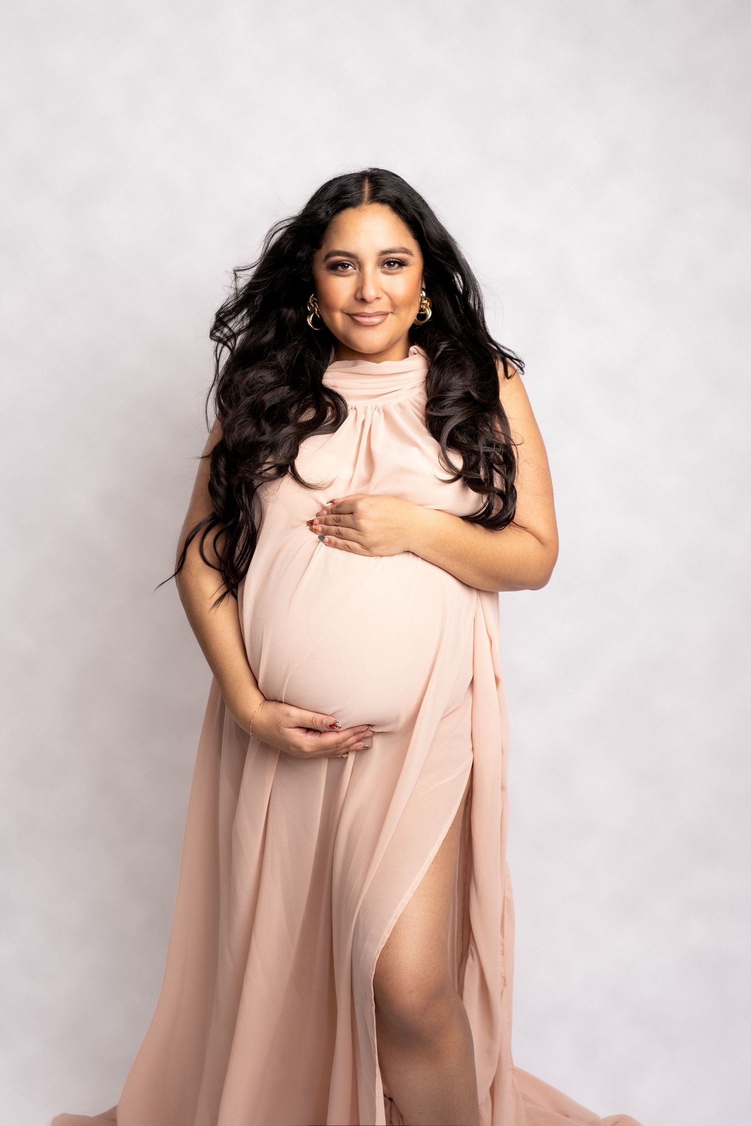 A pregnant woman in a flowing blush pink dress poses elegantly against a light gray studio backdrop.