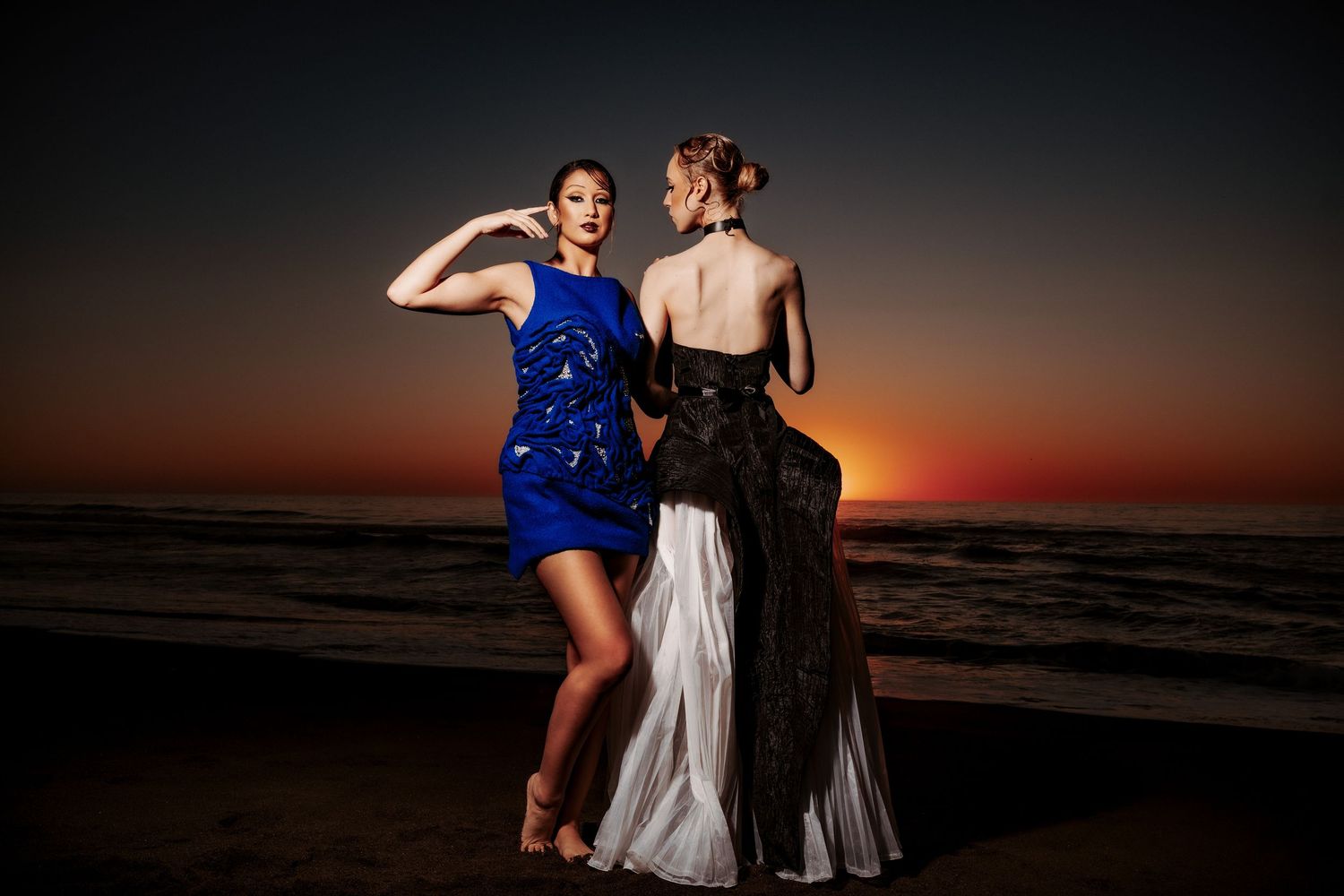 Two friends in elegant gowns dance together at sunset on a beach with dramatic orange sky in the background.