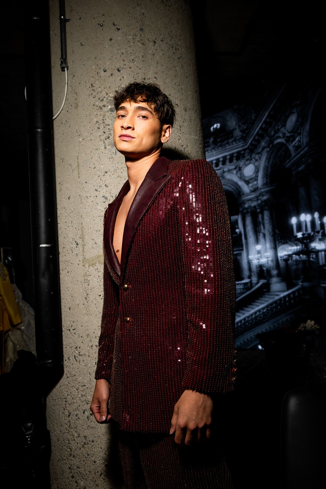 A figure poses against a concrete wall wearing a burgundy sequined blazer with dramatic lighting casting shadows.