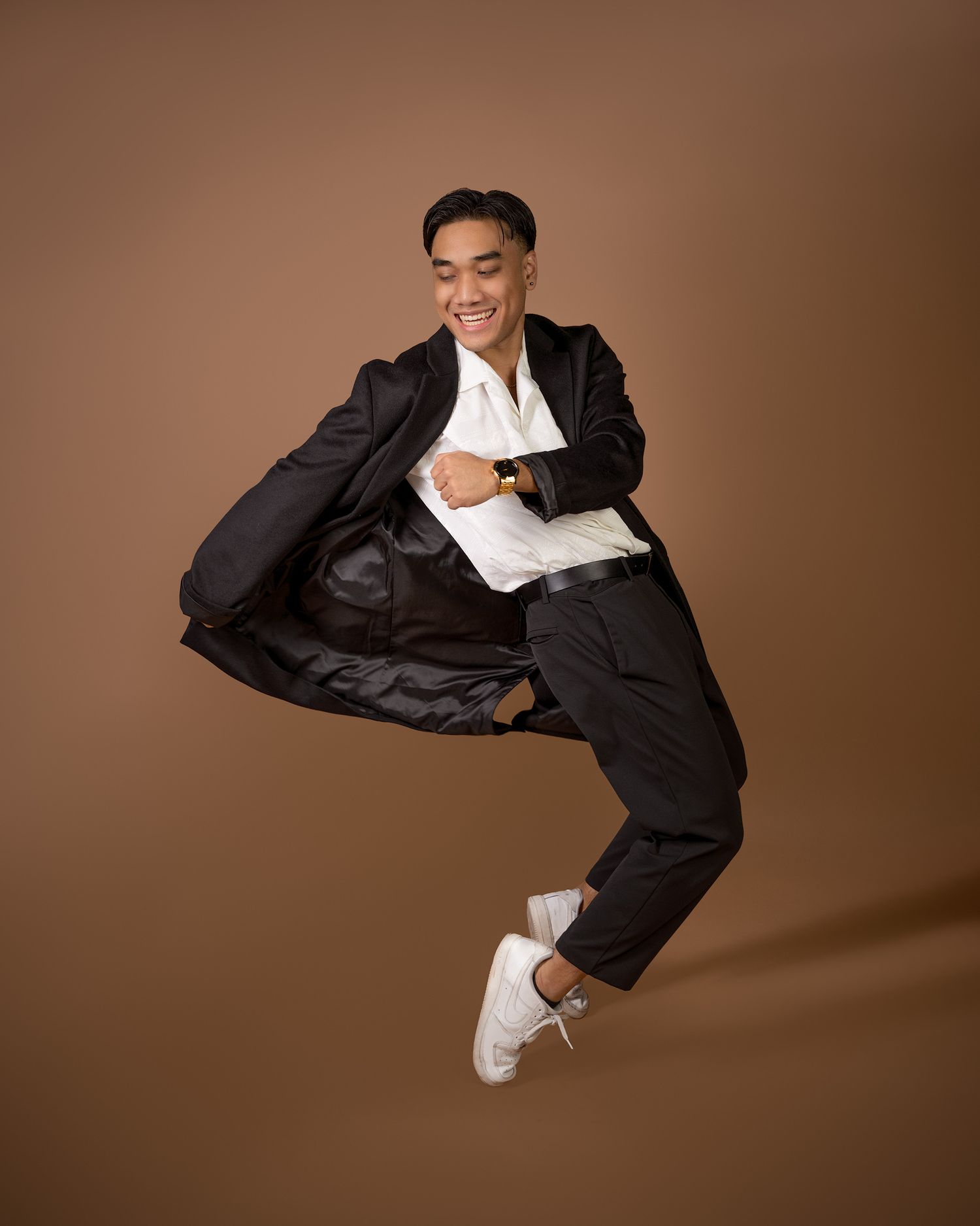 A person in a black suit and white sneakers leaps joyfully against a brown studio background.