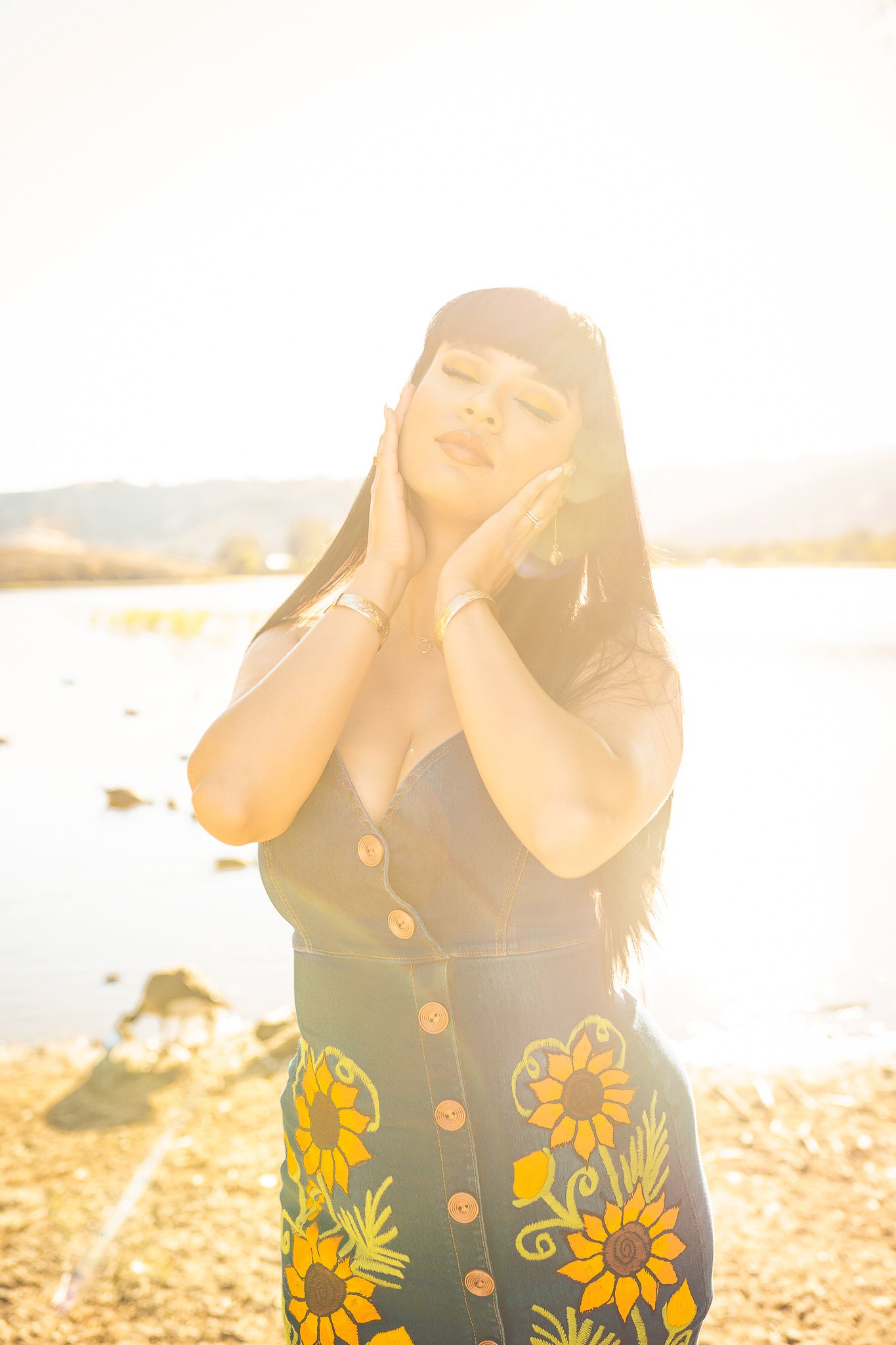 A person in a sunflower-patterned dress poses against golden sunlight by a lake on a warm summer evening.