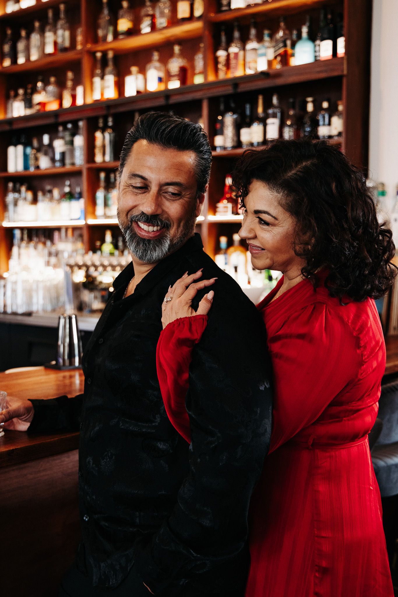 A couple shares a warm moment at a bar with shelves of bottles in the background.
