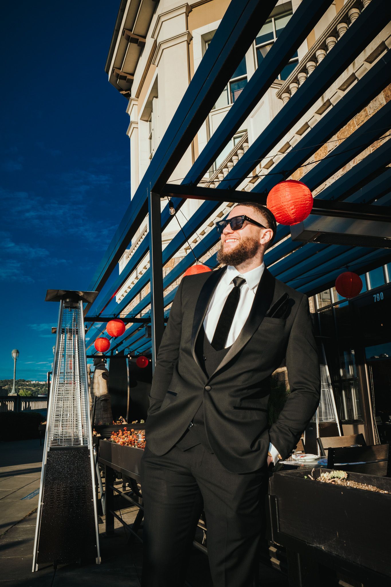 A person in a black suit and sunglasses stands outside a building with red Chinese lanterns and patio heaters.