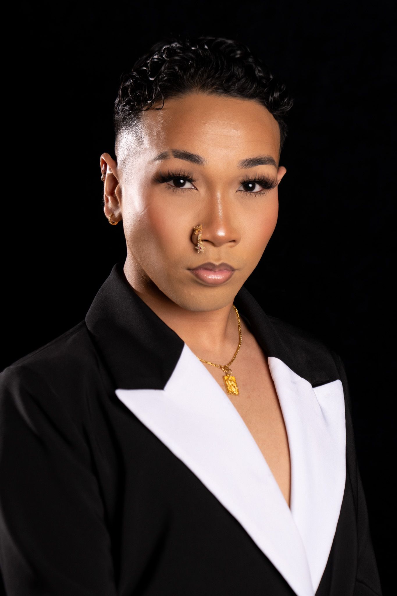 Professional headshot portrait against black background wearing black and white blazer with gold necklace accent.