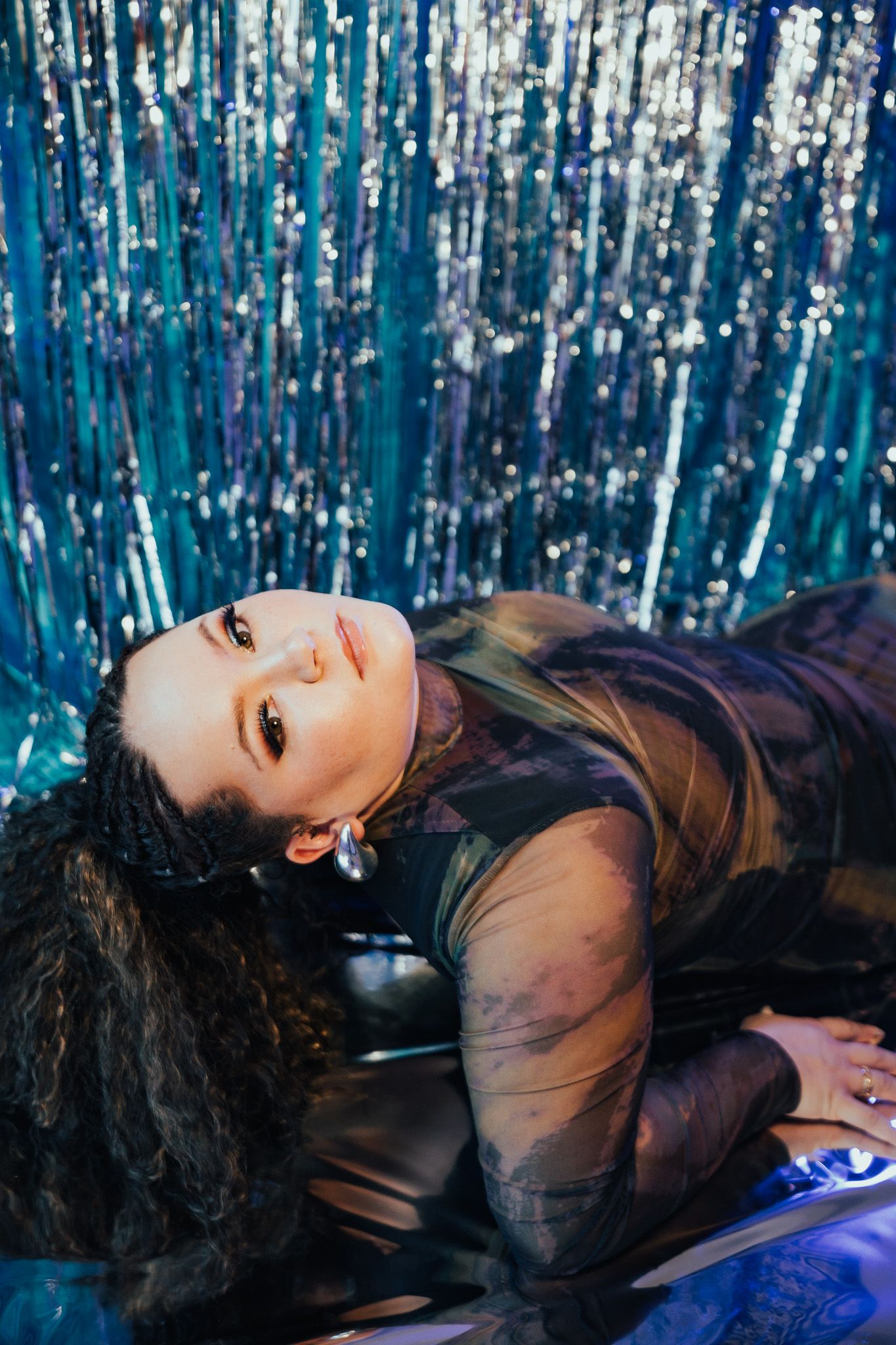 Artistic photo with shimmering blue tinsel backdrop showing figure in sheer black outfit laying down with dramatic lighting.