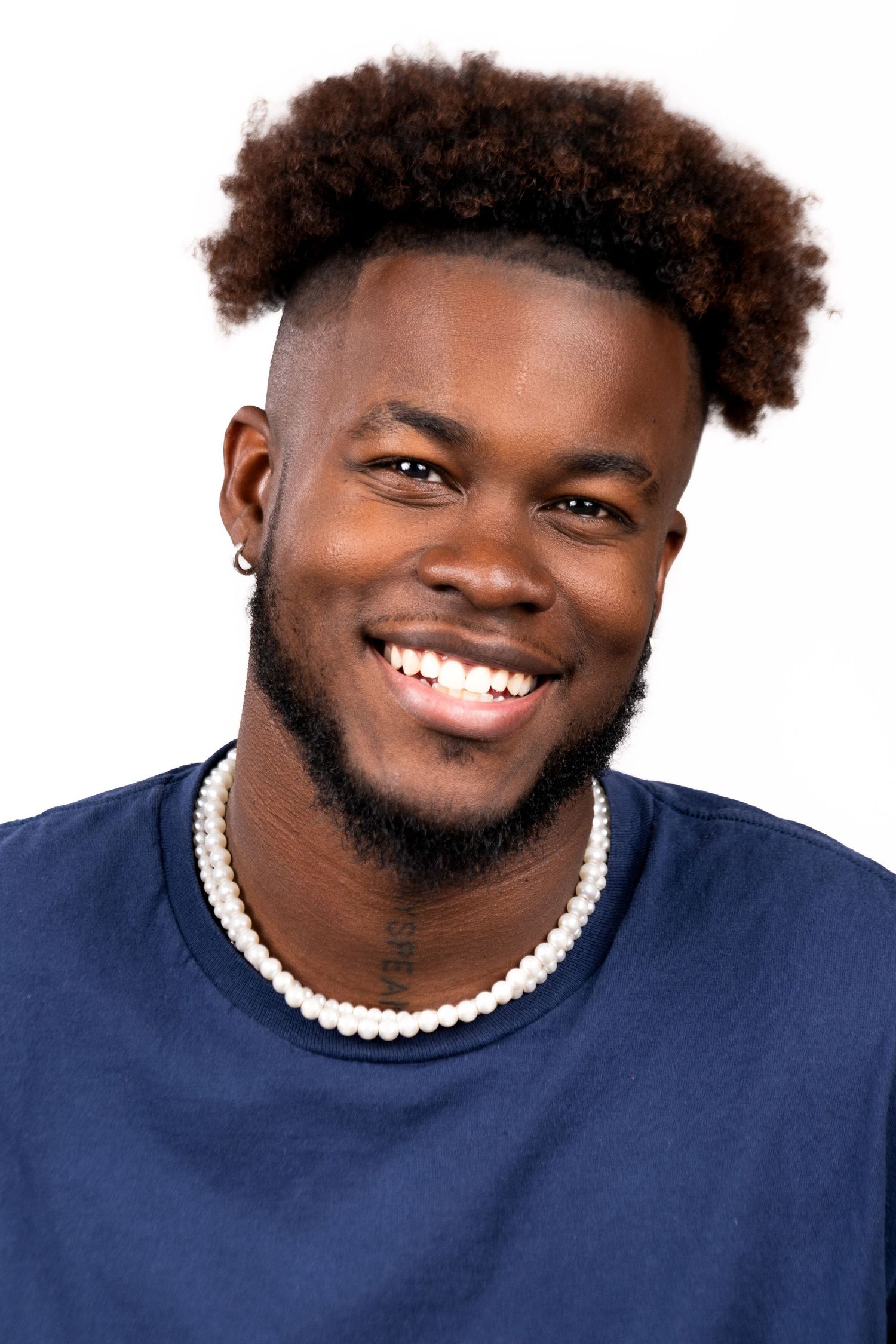 Smiling person wearing a navy blue sweater and pearl necklace against white background with natural curly hair.