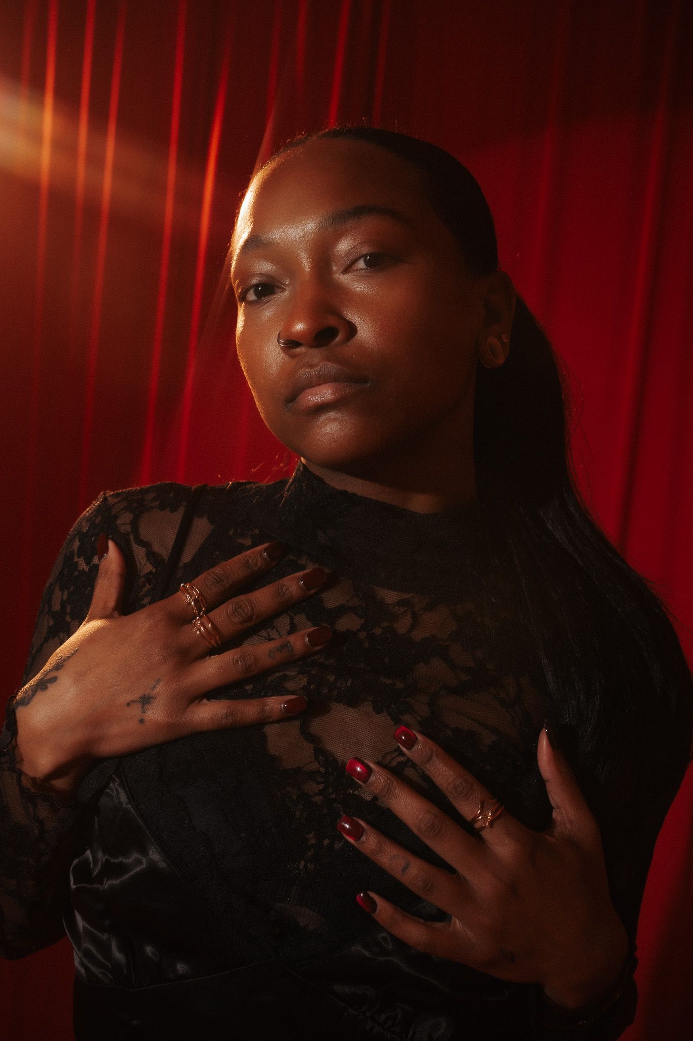 Dramatic portrait against red backdrop shows figure in black lace top with hands crossed over chest and long dark hair.