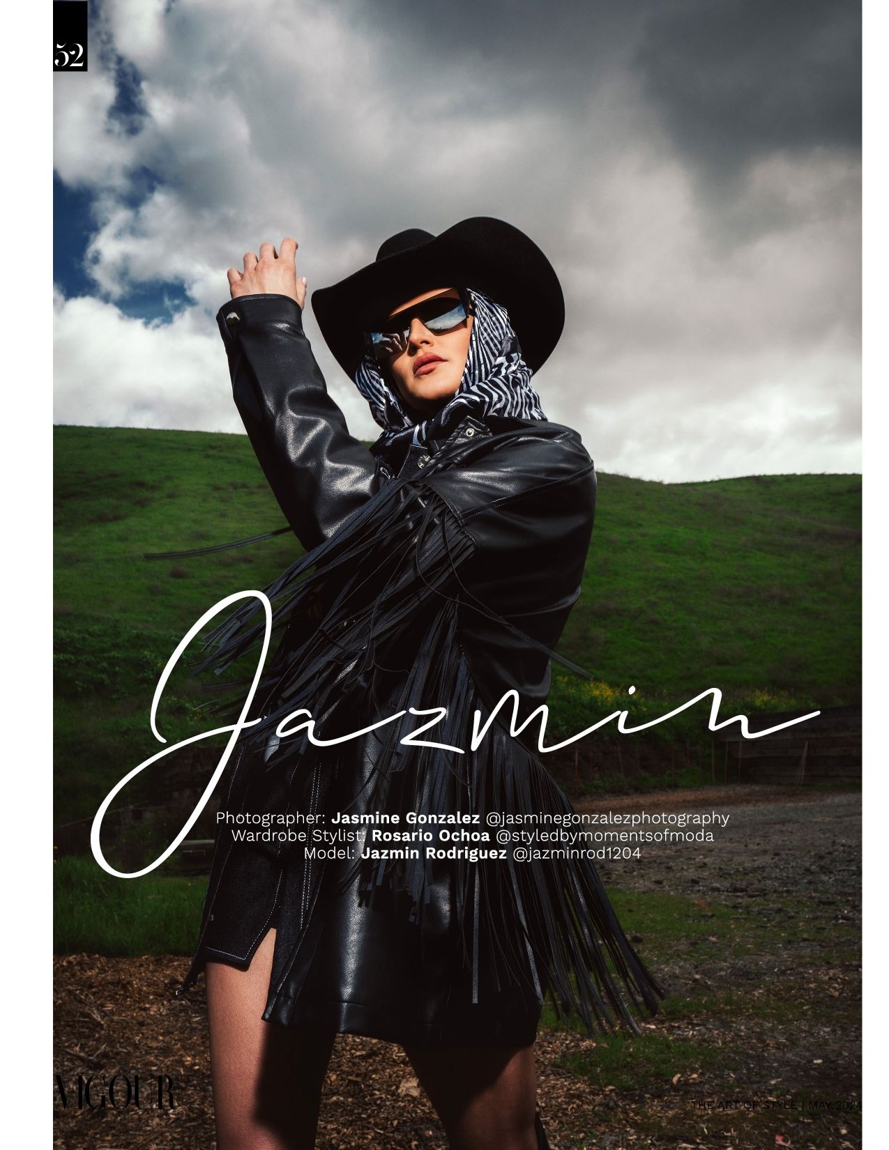 A stylish figure in a black leather jacket, wide-brimmed hat and bandana poses against moody clouds and green hills.