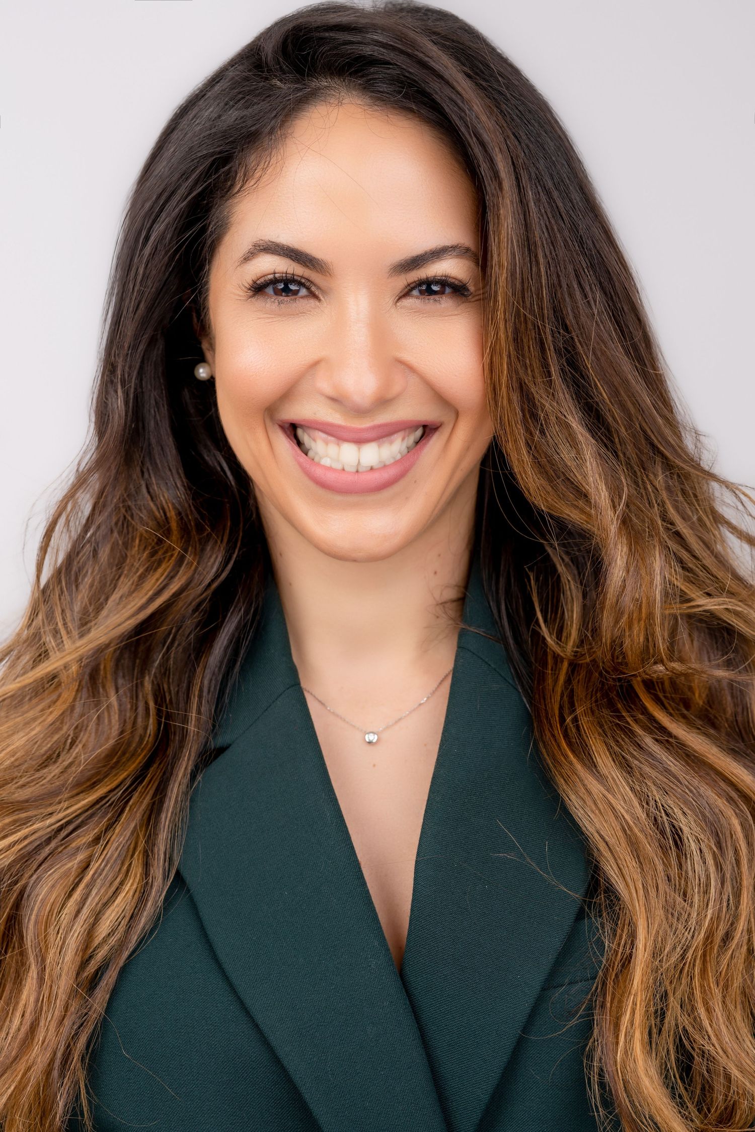 Professional headshot of someone in a dark green blazer with ombre long brown hair and a bright smile.