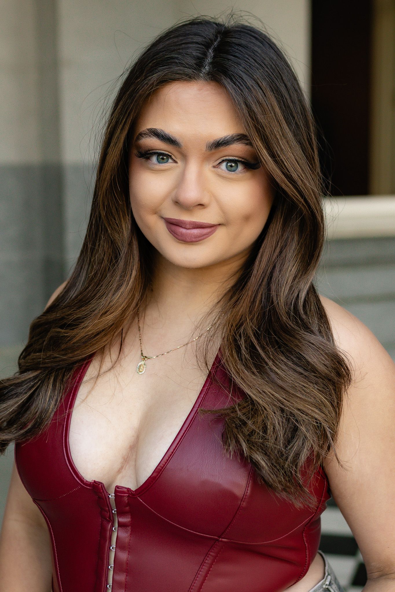 Portrait of someone with long brown wavy hair wearing a burgundy leather top and delicate necklace.