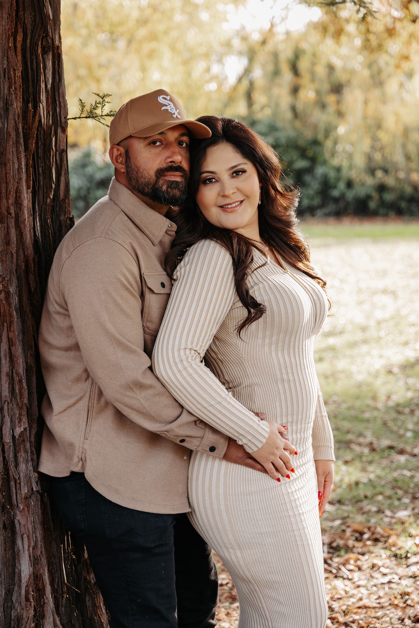 Couple embraces while leaning against tree trunk in autumn setting wearing coordinating neutral-colored outfits.