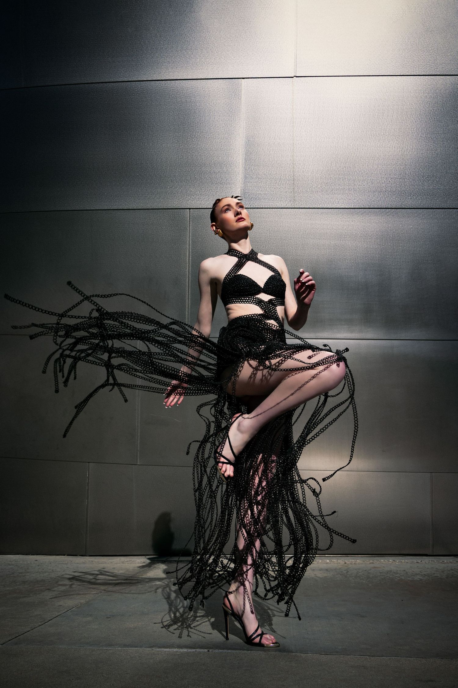Figure in black avant-garde outfit with flowing wire-like tendrils poses dramatically against metallic wall in moody lighting.