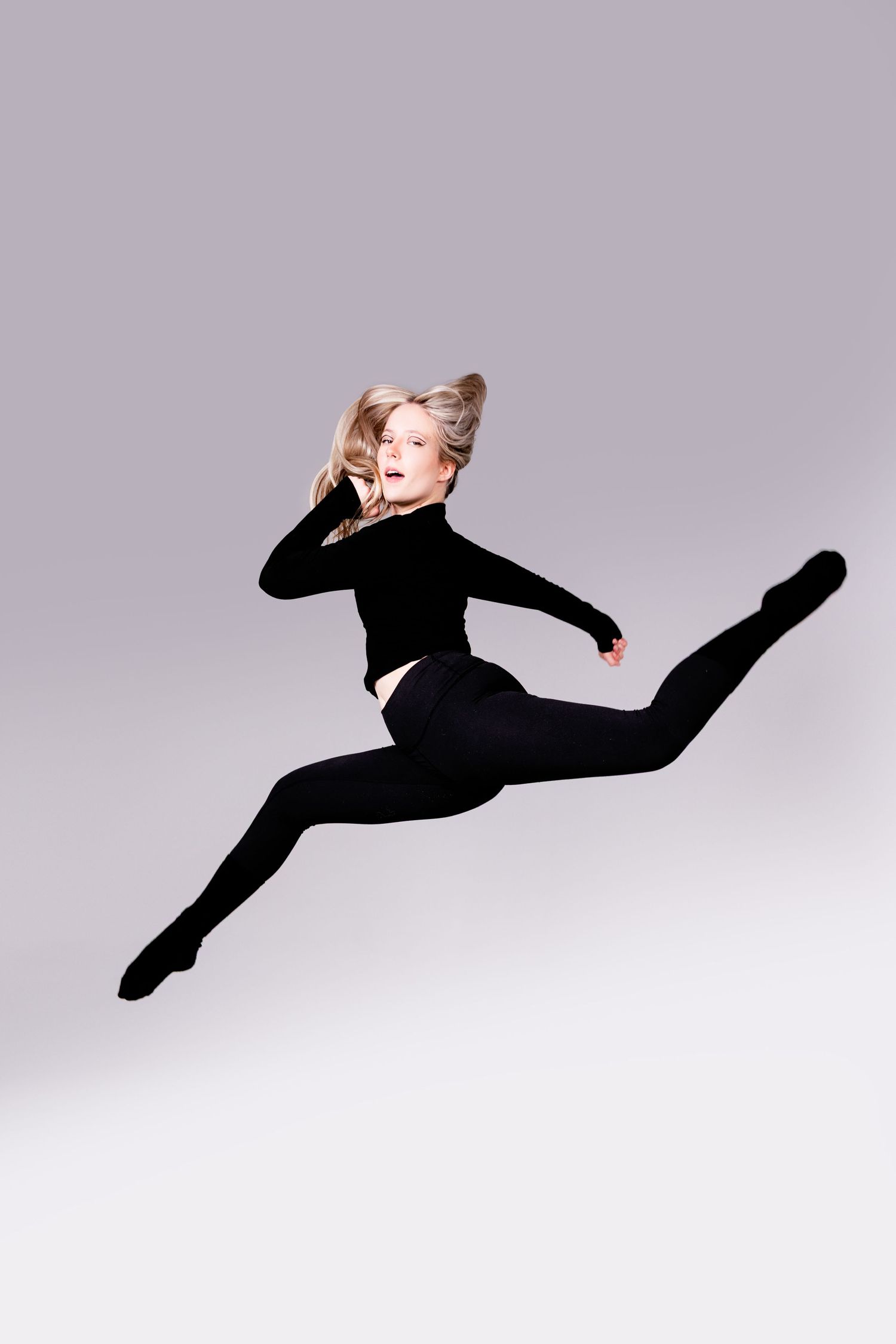 Graceful dancer in black leotard and tights performs a dramatic split leap against a light gray background.