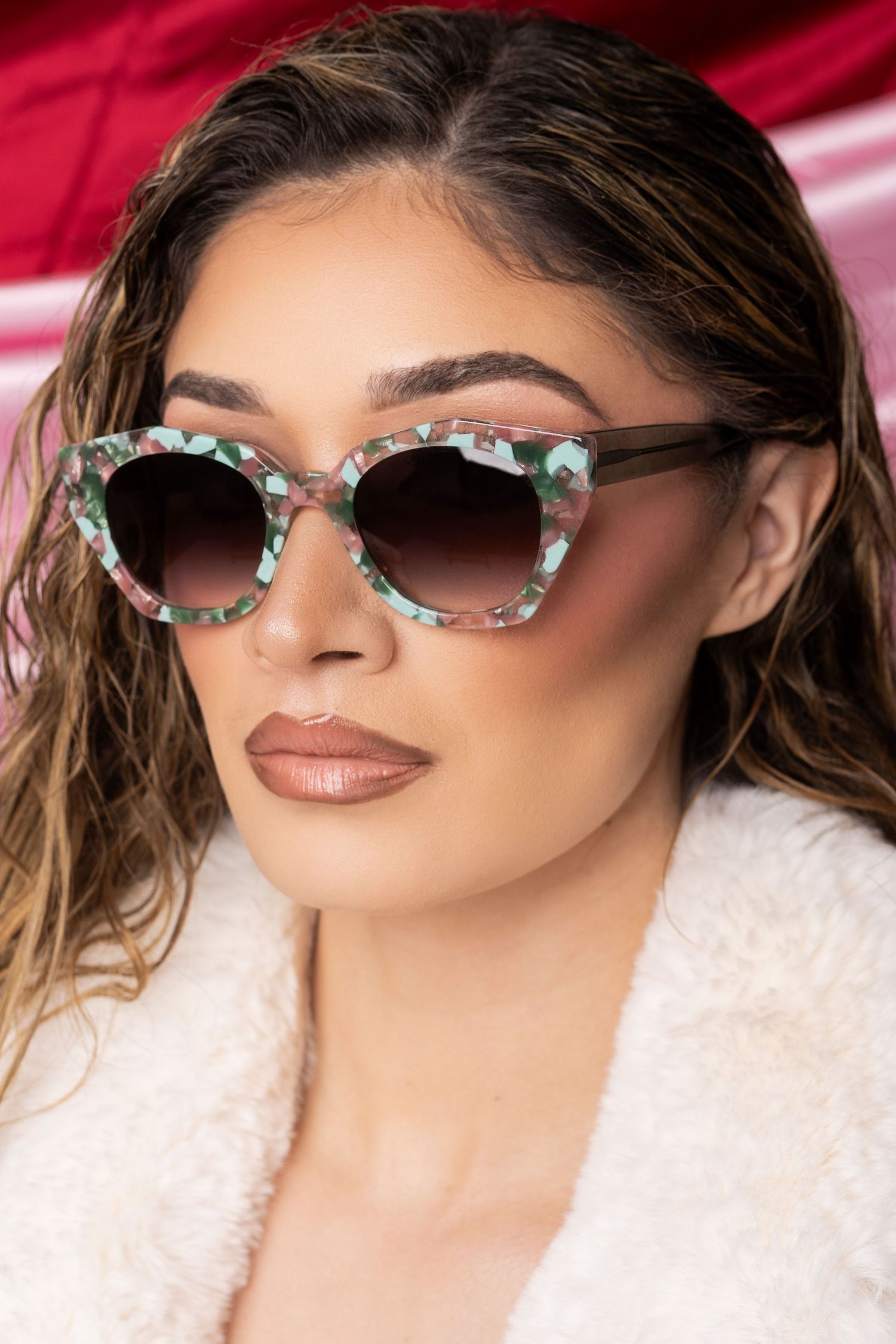 Elegant model wearing floral patterned cat-eye sunglasses and white fuzzy coat against red background.