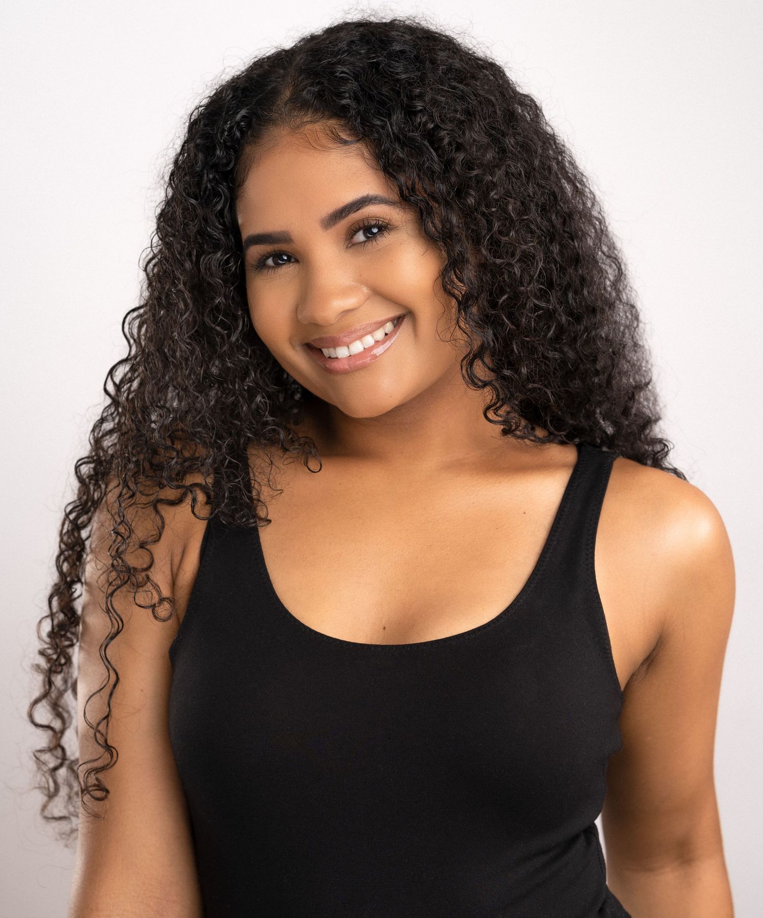Person with long curly dark hair and bright smile wearing a black sleeveless top against white background.