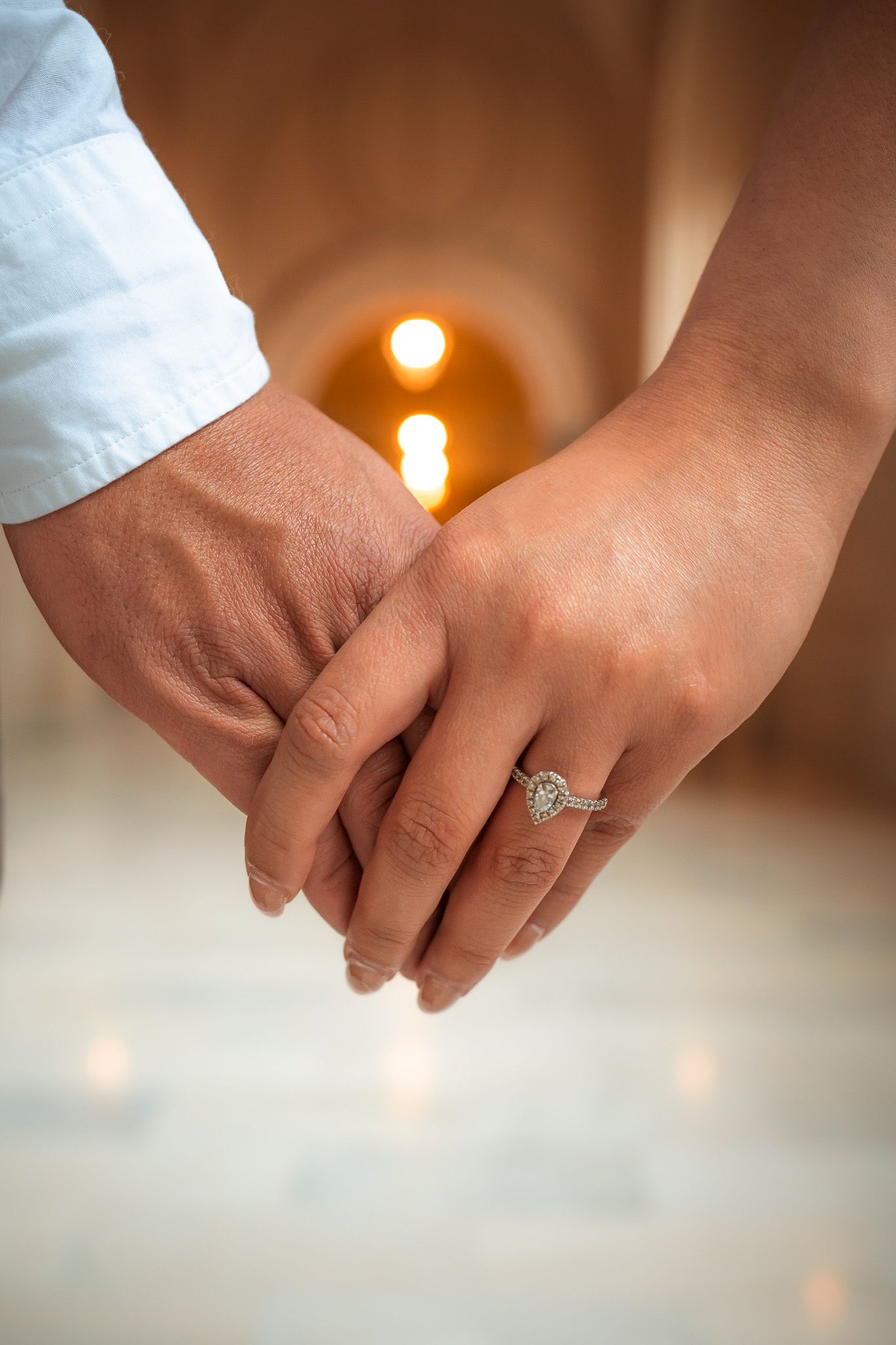 A close-up shot of clasped hands with an engagement ring, illuminated by warm glowing lights in the background.