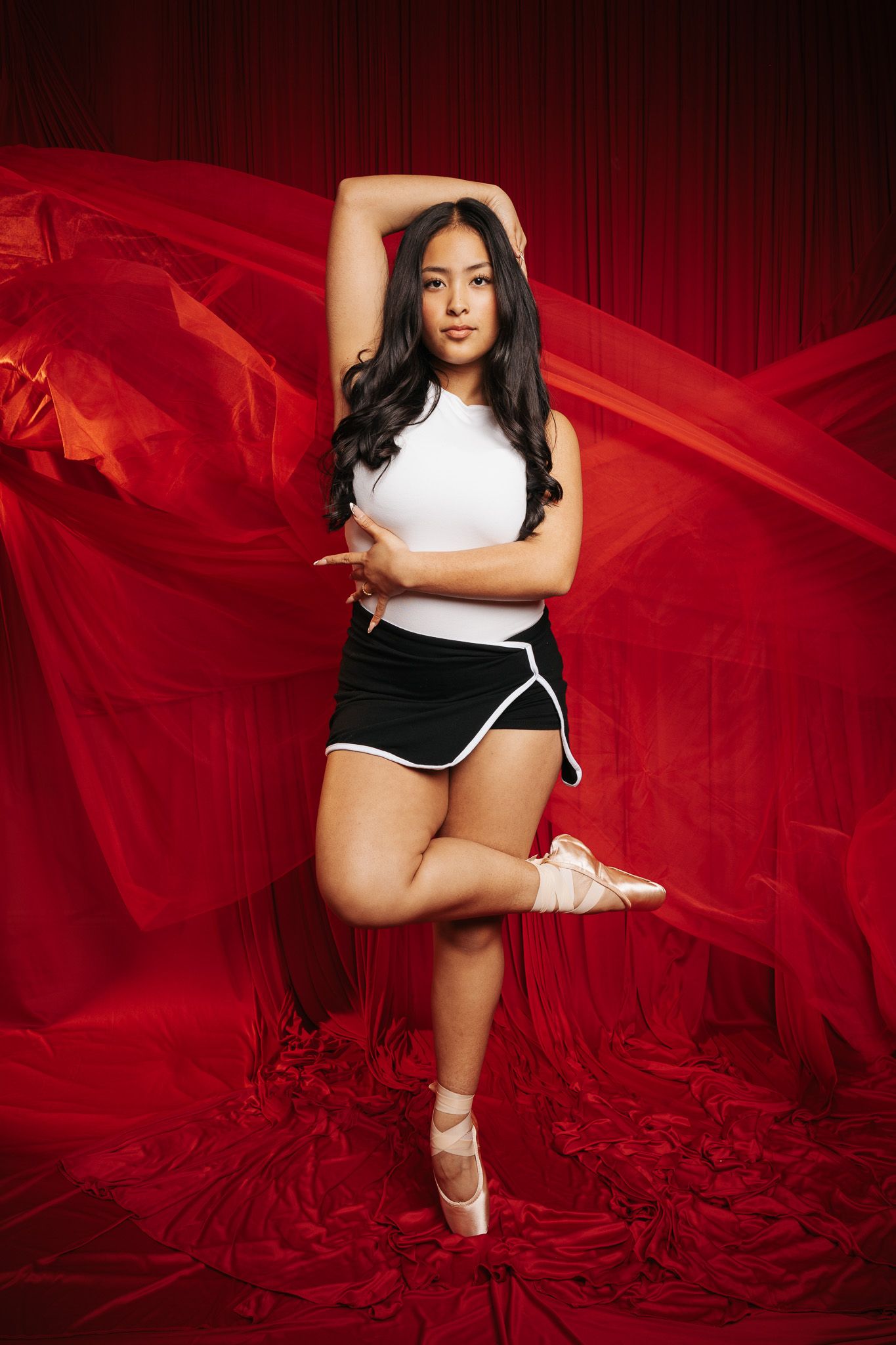 Ballet dancer in white tank top and black shorts poses gracefully against flowing red fabric backdrop.