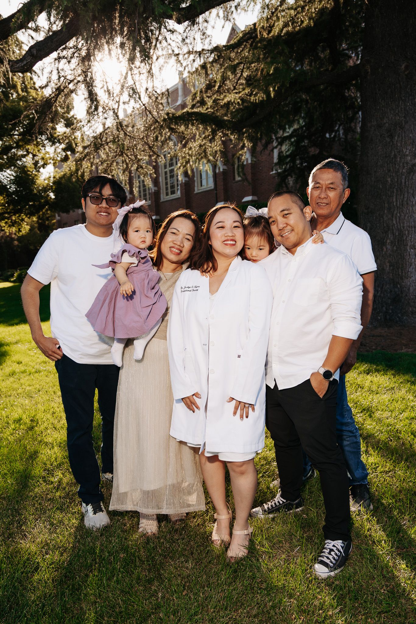 A multi-generational family poses together outdoors under a large tree on a sunny day.