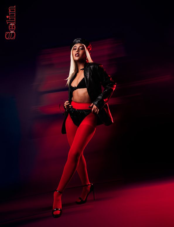 Fashion photo of a model in red tights and black leather jacket striking a dramatic pose against dark red lighting.
