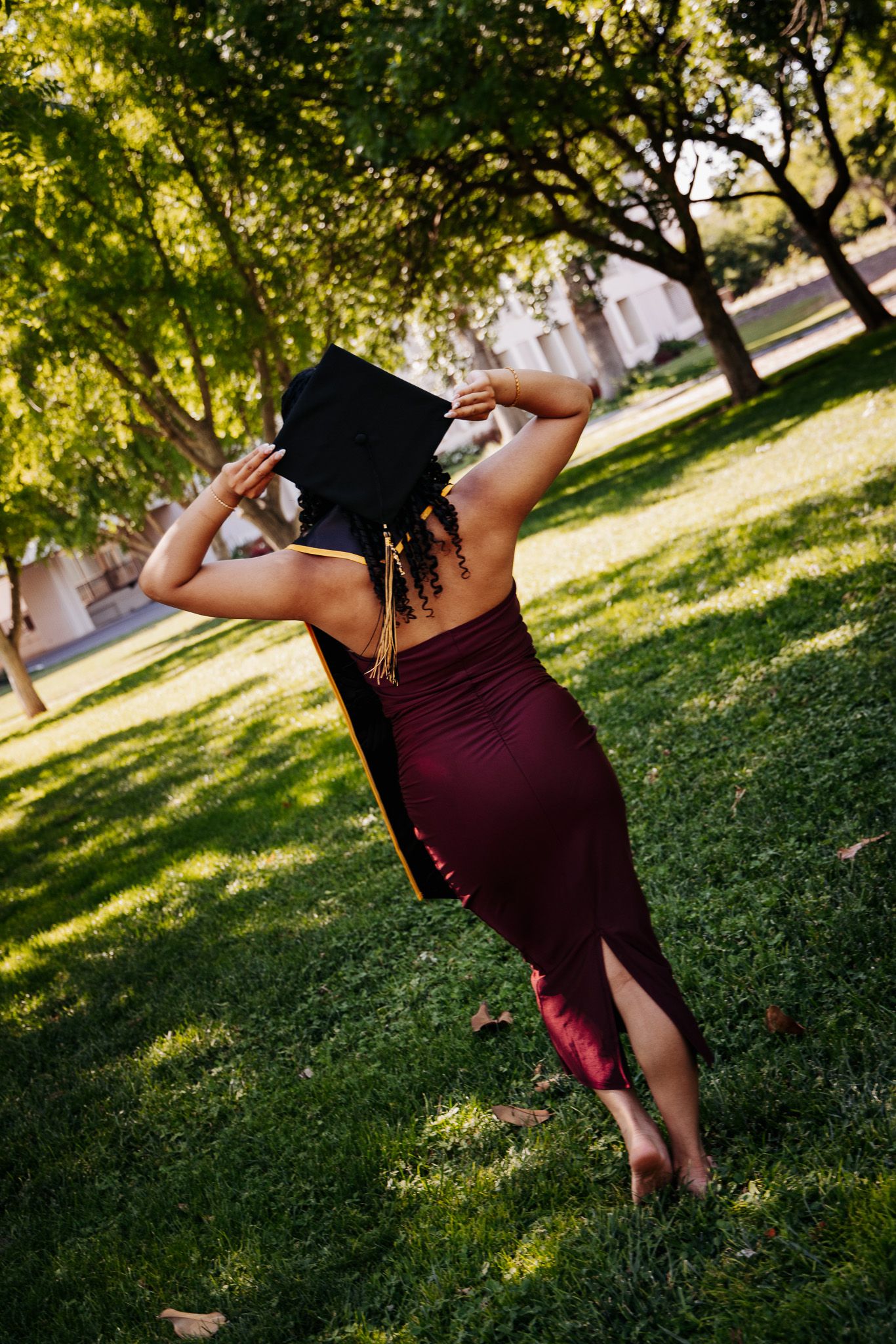 A graduate in a burgundy dress holds their mortarboard cap while standing on grass under leafy trees on a sunny day.