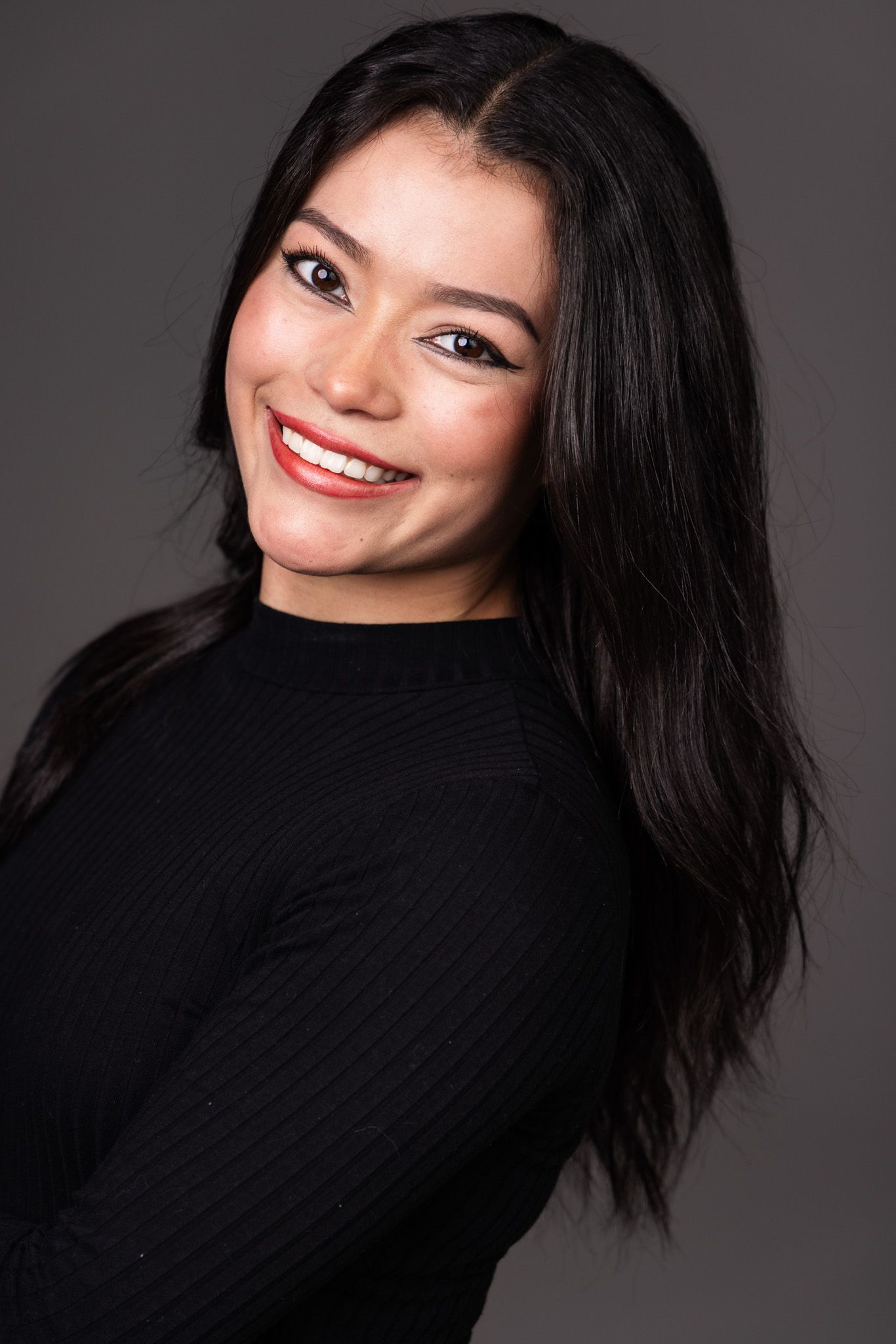 Portrait of a smiling woman with long black hair wearing a black sweater against a dark gray background.