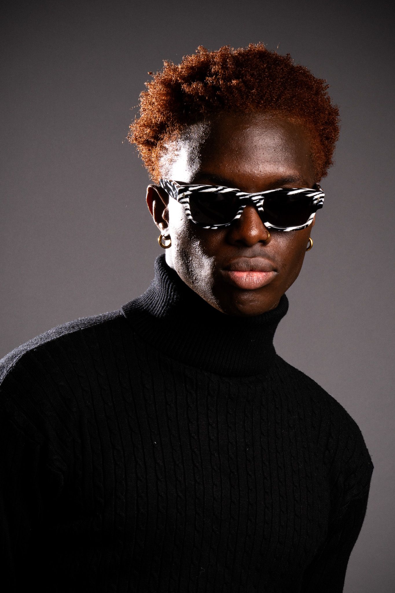 A fashion portrait showing a person wearing stylish sunglasses and a black turtleneck sweater against a dark background.