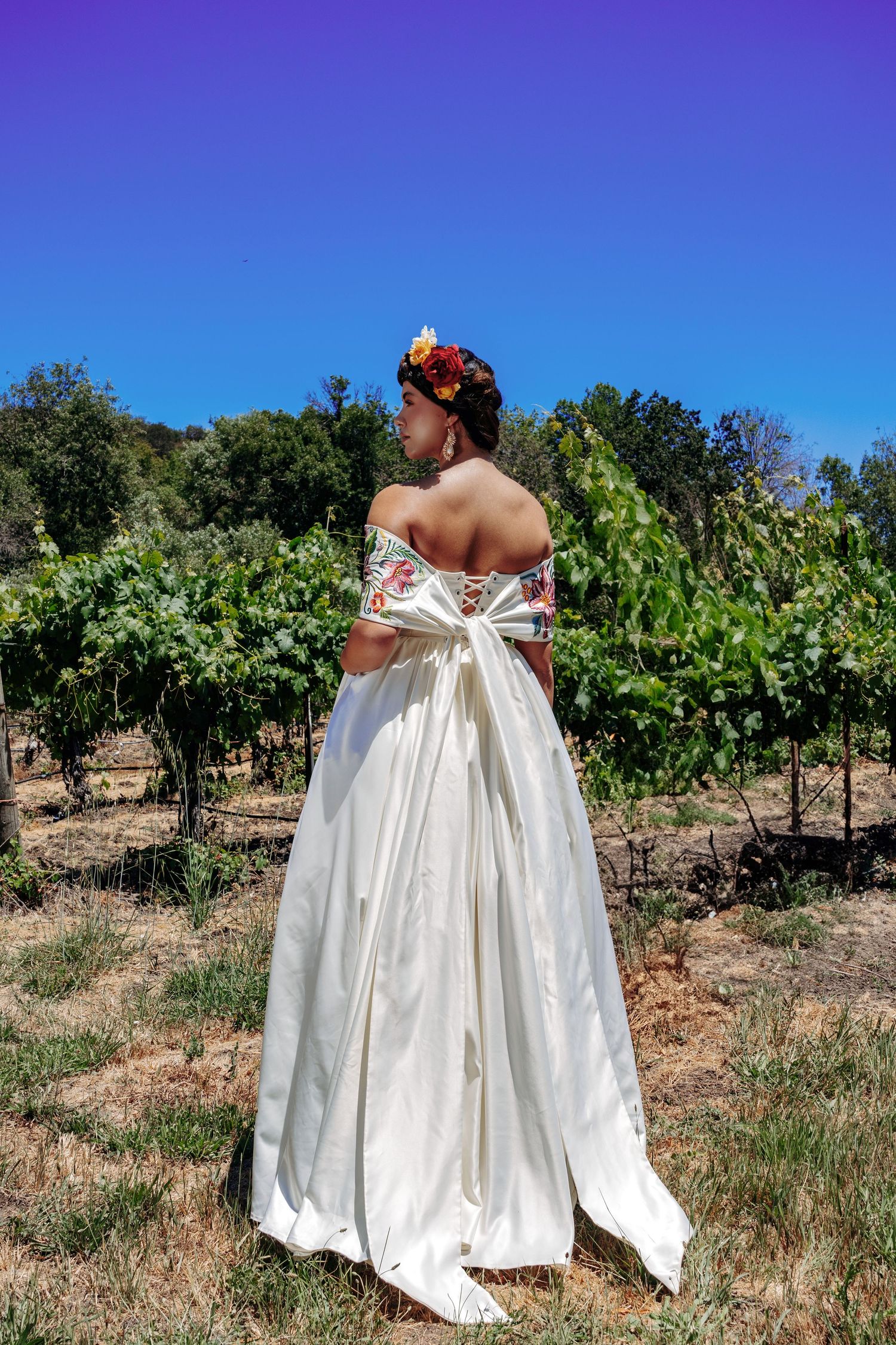 A woman in a white off-shoulder wedding dress stands in a vineyard with floral headpiece under bright blue sky.