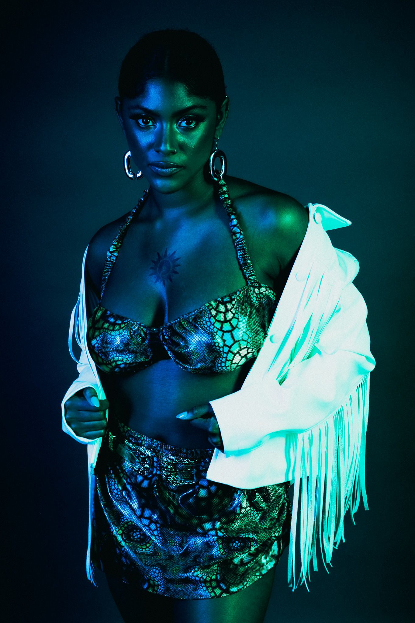 Fashion model wearing sparkly lingerie and white fringe jacket poses in dramatic blue and green lighting.